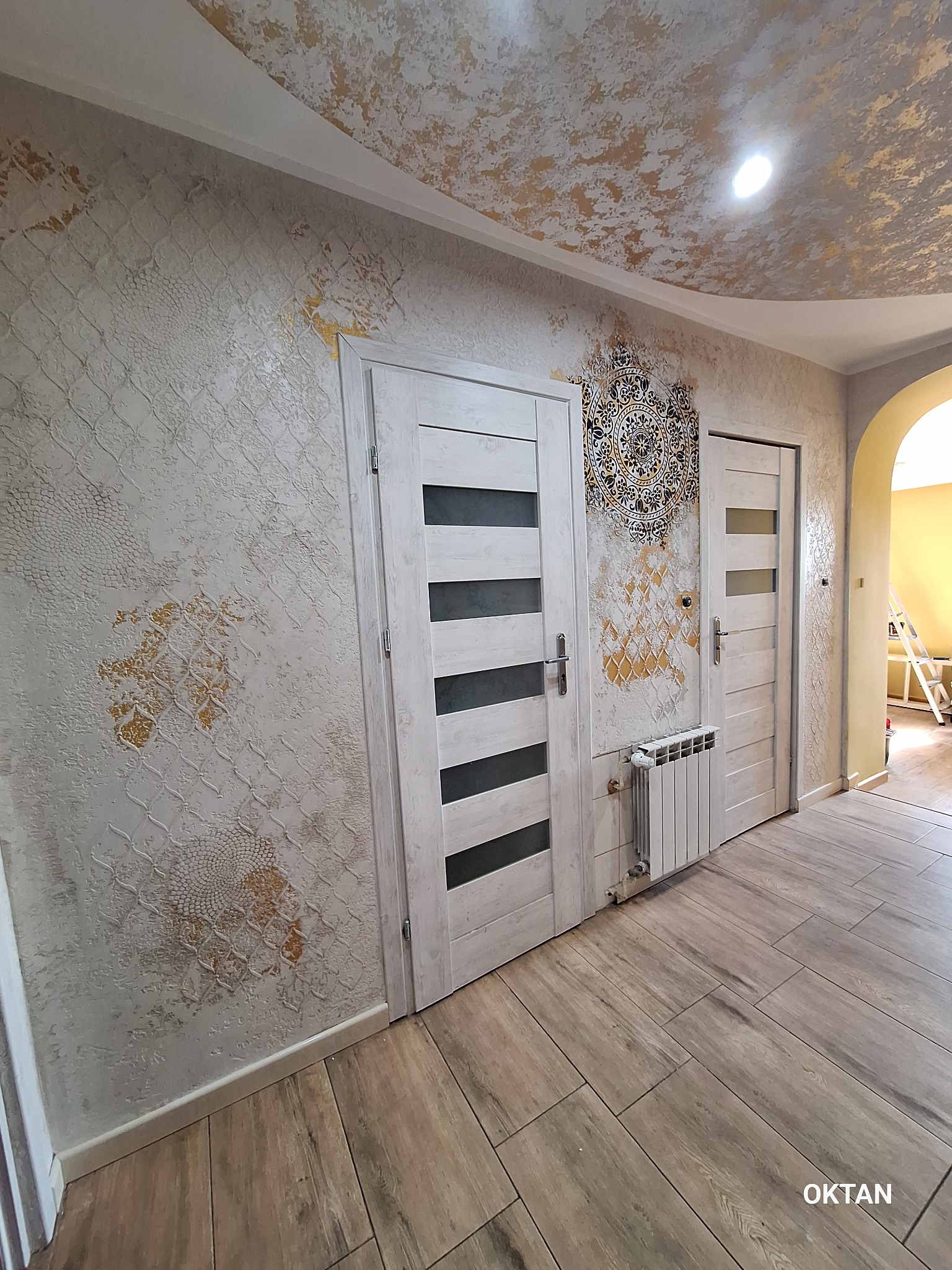 Wnętrze mieszkania po remoncie: dekoracyjne malowanie ścian z efektem struktury, złote ornamenty, drewnopodobne panele podłogowe, białe drzwi z przeszkleniami i grzejnik.