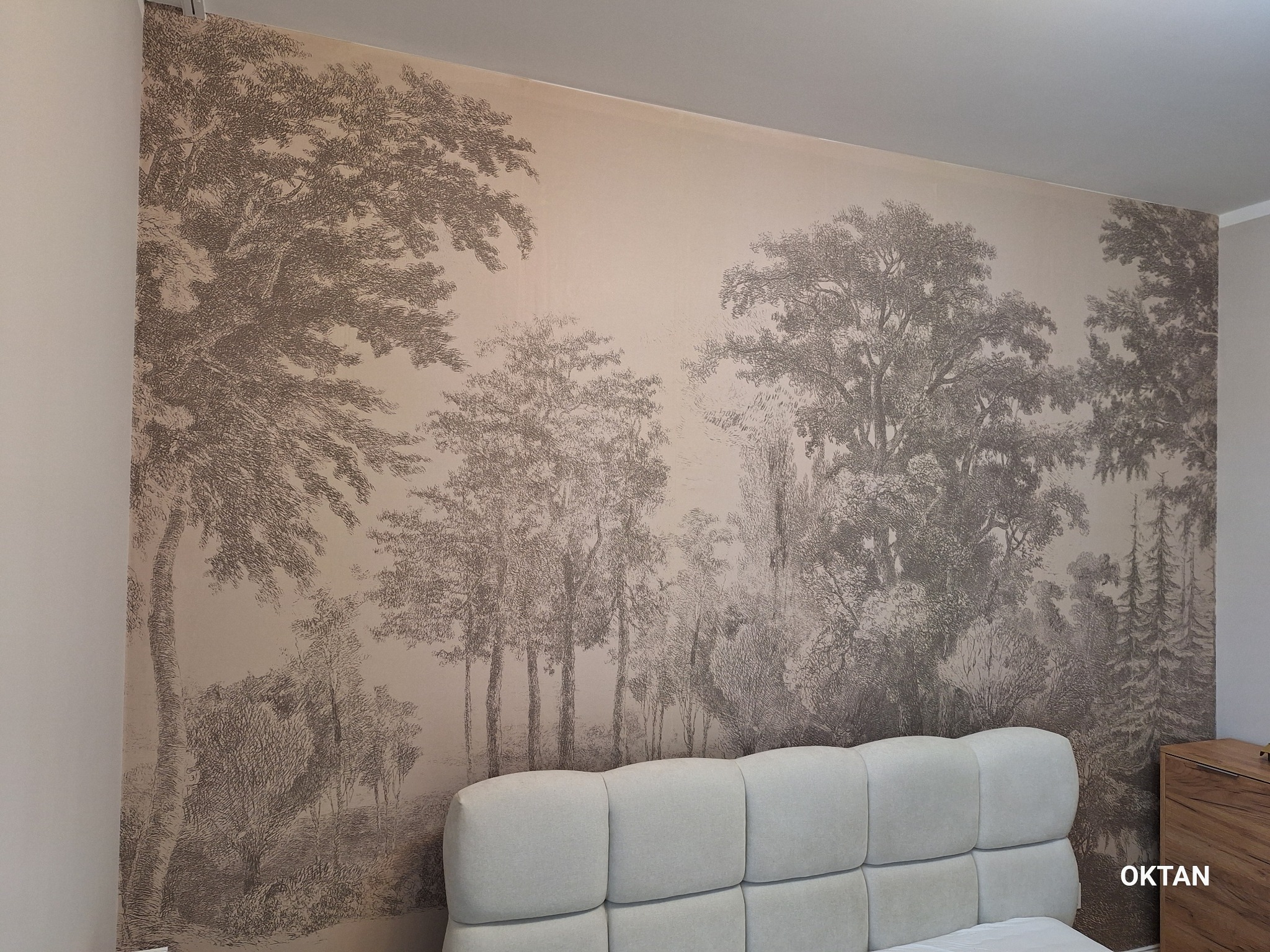 Ściana z tapetą w stylu retro, przedstawiającą leśny krajobraz w odcieniach sepii. Tapeta pokrywa całą ścianę nad łóżkiem z jasnym zagłówkiem.
