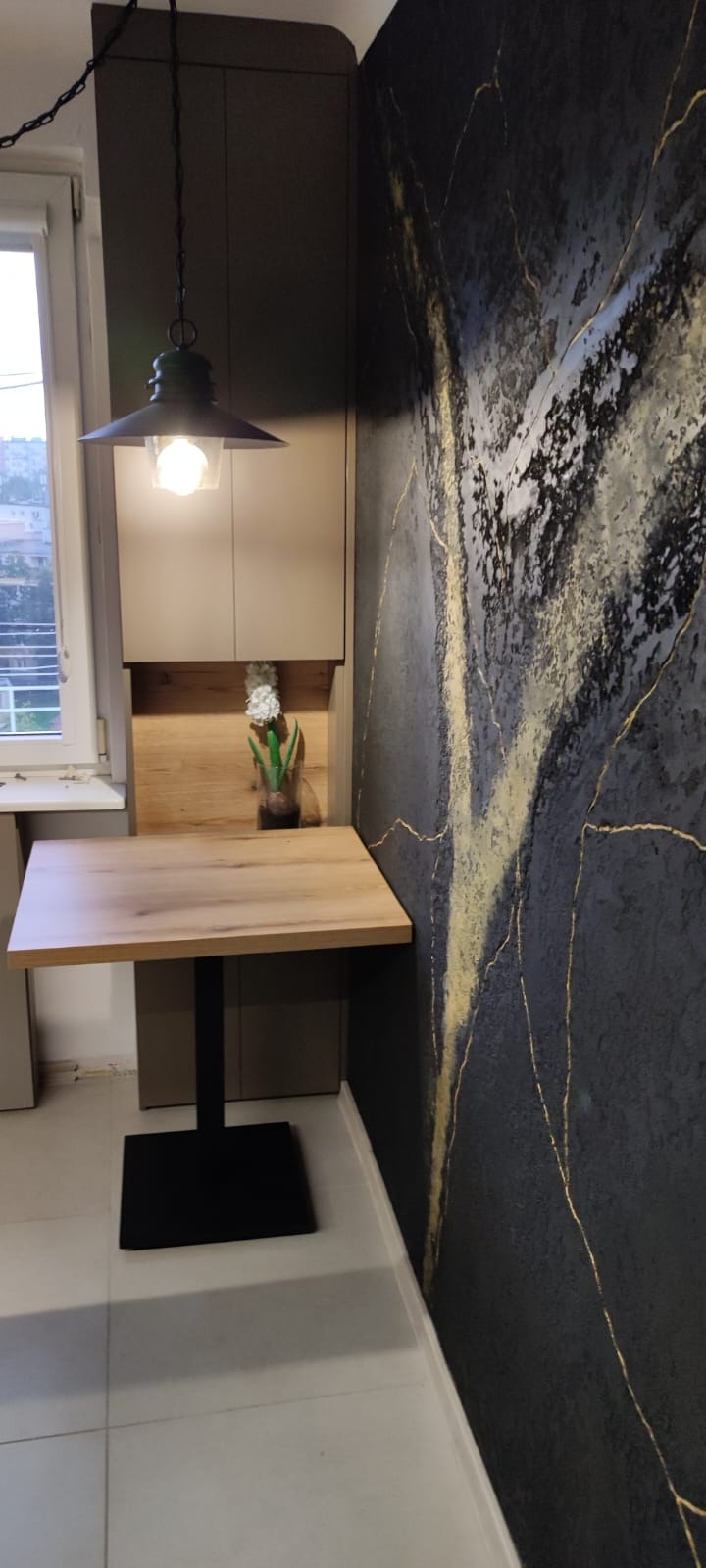 Kompaktowy stół z blatem imitującym drewno, czarna noga, designerska ściana w odcieniach szarości z złotymi akcentami, wisząca lampa w stylu industrialnym, kwiaty w wazonie.