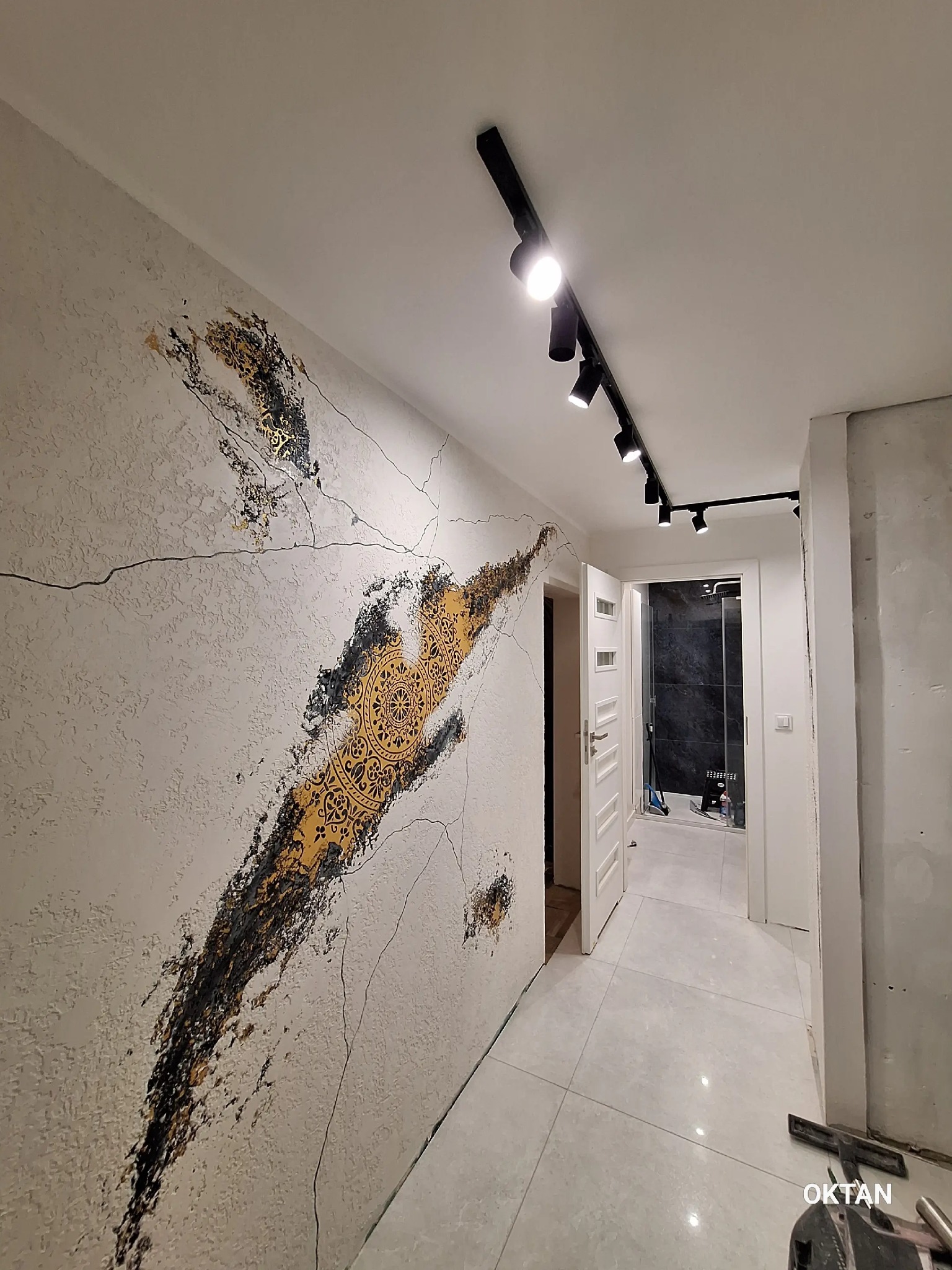 Korytarz z dekoracyjną ścianą w odcieniach bieli, złota i czerni, oświetlony reflektorami na szynie. Widoczne drzwi do łazienki i fragment podłogi z płytek.