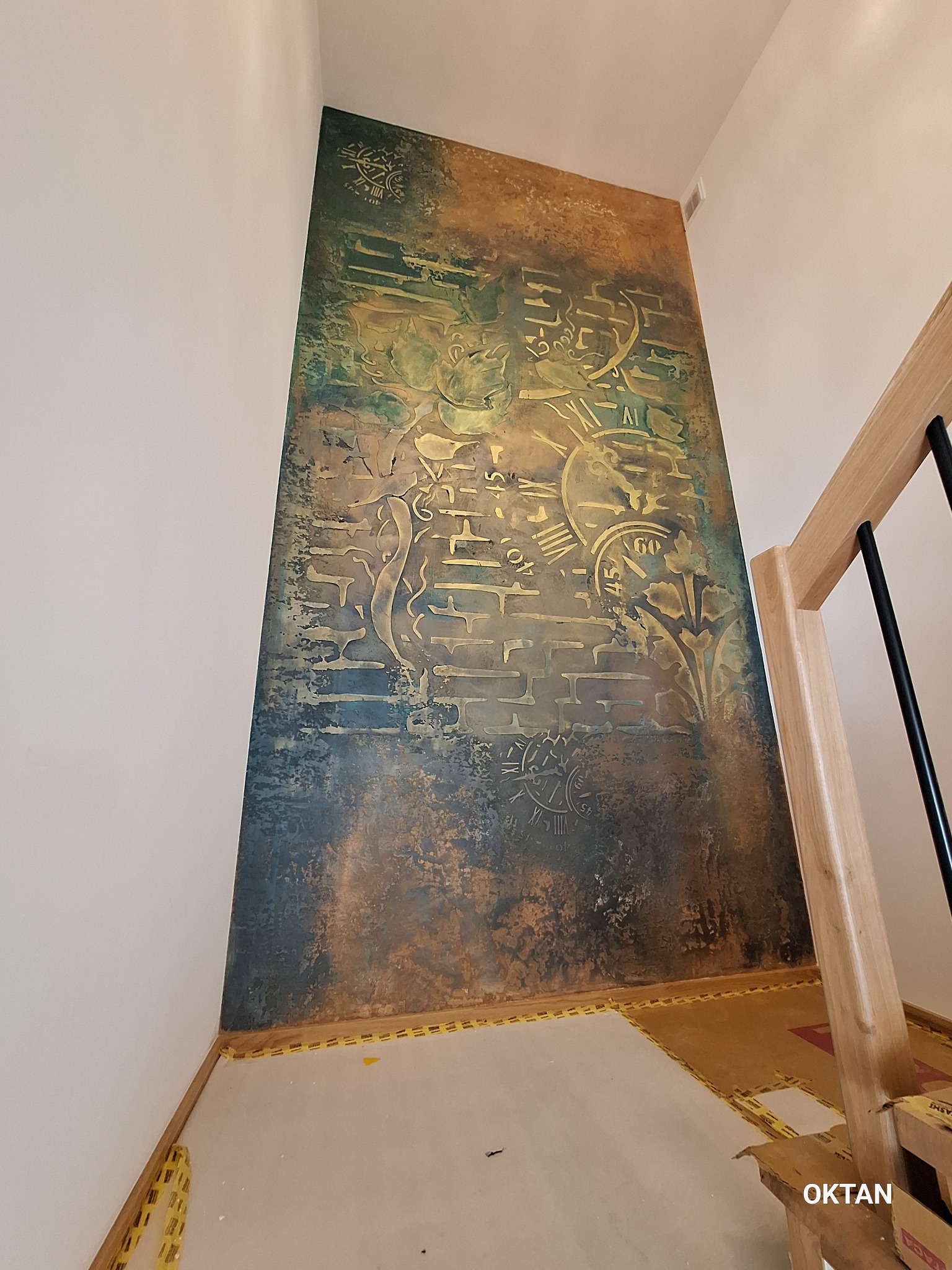 Dekoracyjna ściana z efektem rdzy i motywem zegara, widoczna z dołu, z fragmentem drewnianej balustrady schodów. Żółta taśma malarska zabezpiecza podłogę.