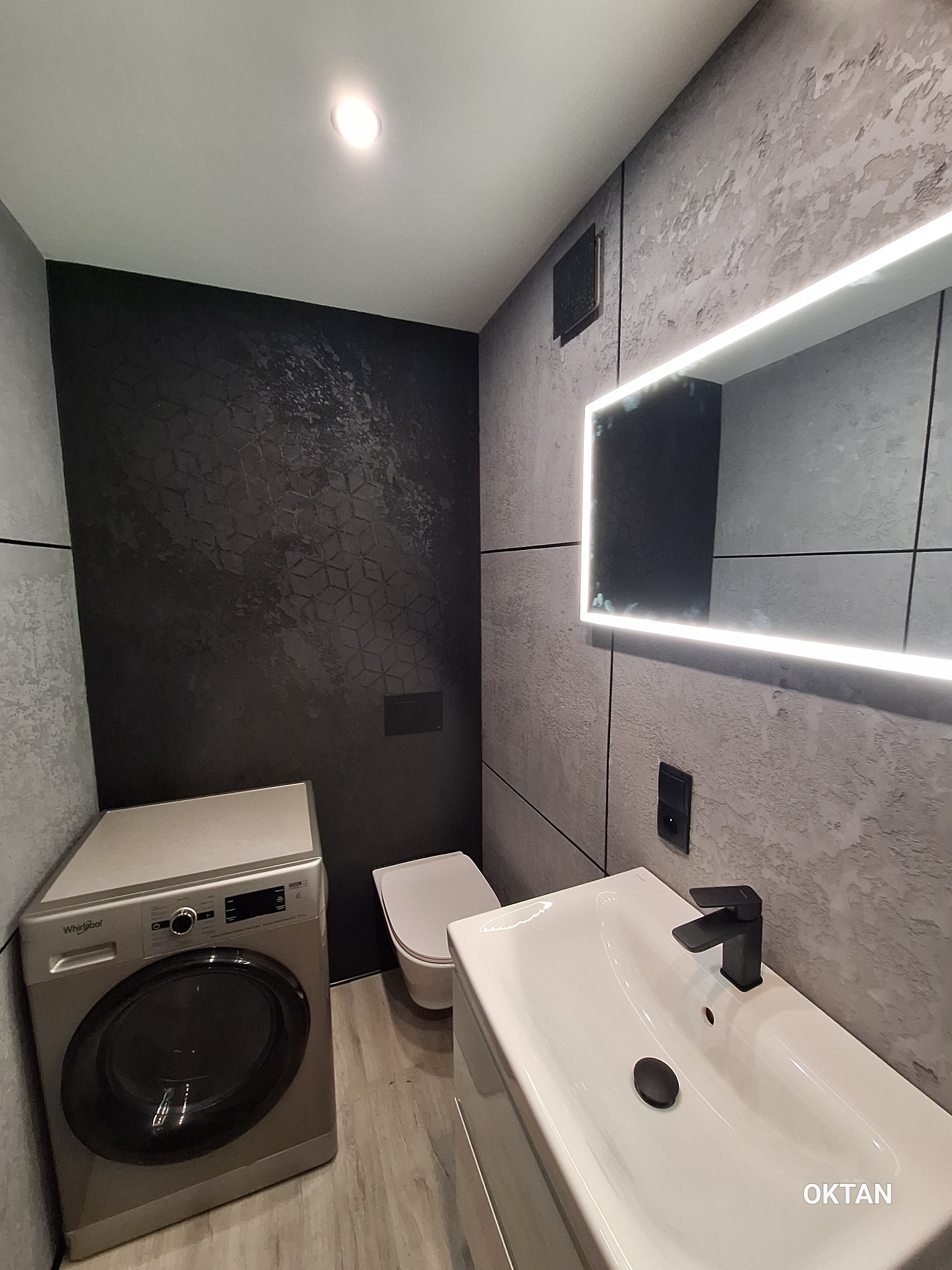 Wykończona łazienka w szarościach z podświetlanym lustrem, pralką i umywalką. Na ścianie dekoracyjna, czarna tapeta z geometrycznym wzorem. Nowoczesny design.