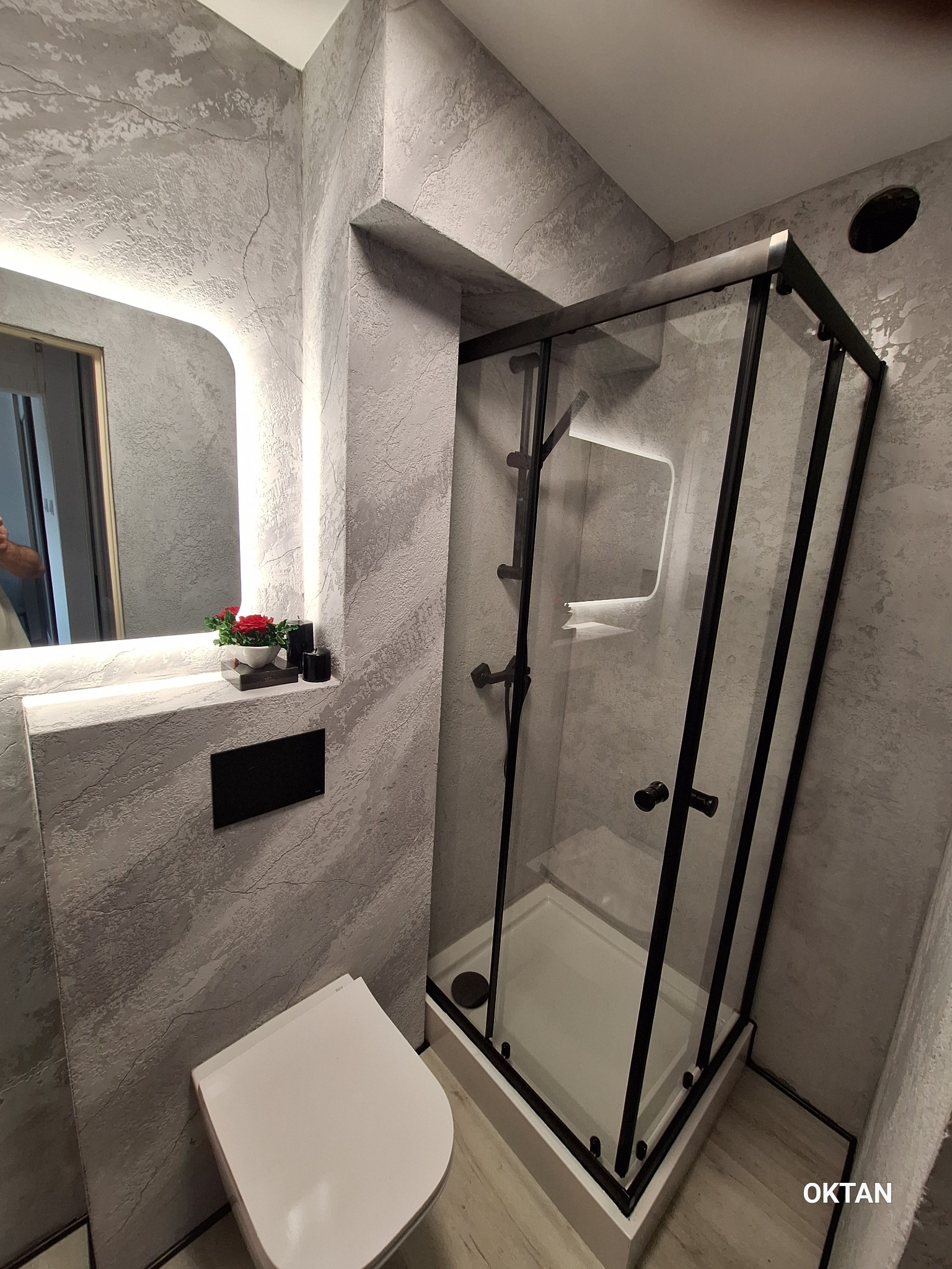 Nowoczesna łazienka z kabiną prysznicową typu walk-in, toaletą i lustrem z podświetleniem LED. Ściany pokryte dekoracyjną szarą strukturą. Minimalistyczny design.