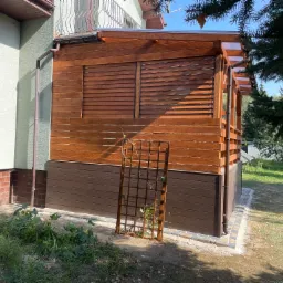 Zadaszona konstrukcja z drewnianymi żaluzjami, przylegająca do budynku, z drewnianą kratką na pnącza w pobliżu podstawy. Widoczny fragment rynny i fragment ogrodu.