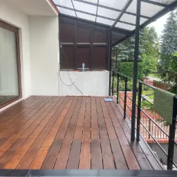 Zadaszony taras z drewnianą podłogą w trakcie budowy, widoczne narzędzia i materiały budowlane, czarna metalowa balustrada, widok na zieleń ogrodu