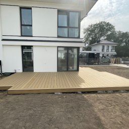 TarasDesign - Jasny taras z desek kompozytowych przy nowoczesnym domu z dużymi oknami. Widoczny fragment ogrodu i drabina oparta o ścianę budynku.