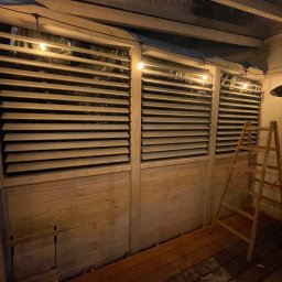 TarasDesign - Drewniana konstrukcja z żaluzjami, oświetlona girlandą żarówek, z drewnianą podłogą i drabiną. Widoczne elementy montażowe i folia zabezpieczająca.