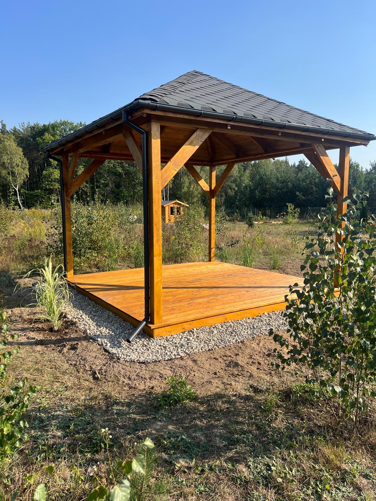 Nowoczesna, drewniana konstrukcja z dachem krytym gontem bitumicznym, otoczona kamiennym obrzeżem i zielenią, w tle widoczny mały domek.