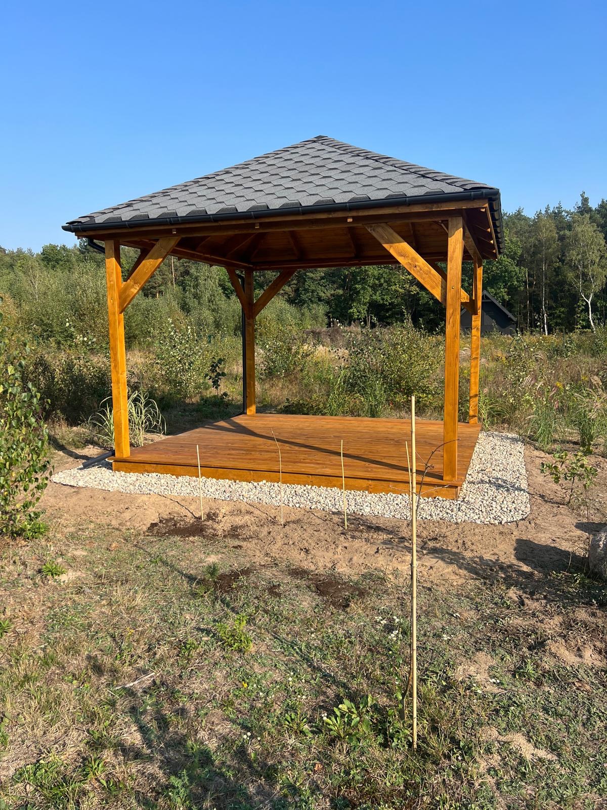 Nowoczesna, otwarta konstrukcja z drewnianymi słupami i szarym, spadzistym dachem z gontów bitumicznych, otoczona kamiennym obramowaniem i młodymi drzewkami, na tle zieleni.