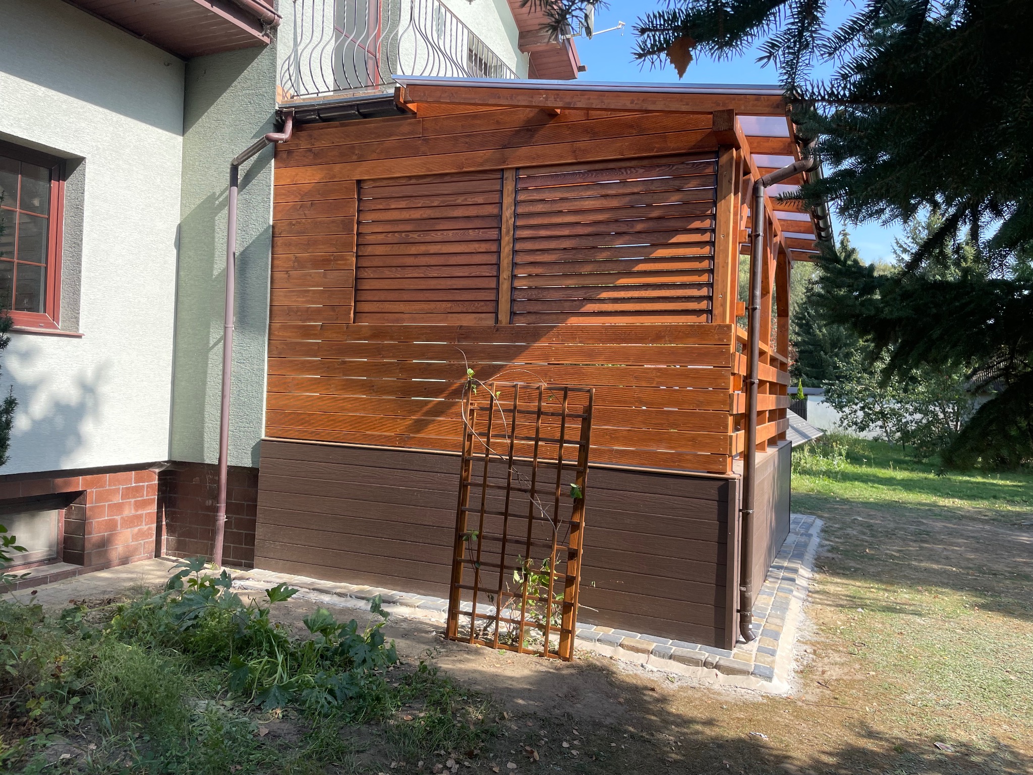 Zadaszona konstrukcja z drewnianymi żaluzjami, przylegająca do budynku, z drewnianą kratką na pnącza w pobliżu podstawy. Widoczny fragment rynny i fragment ogrodu.