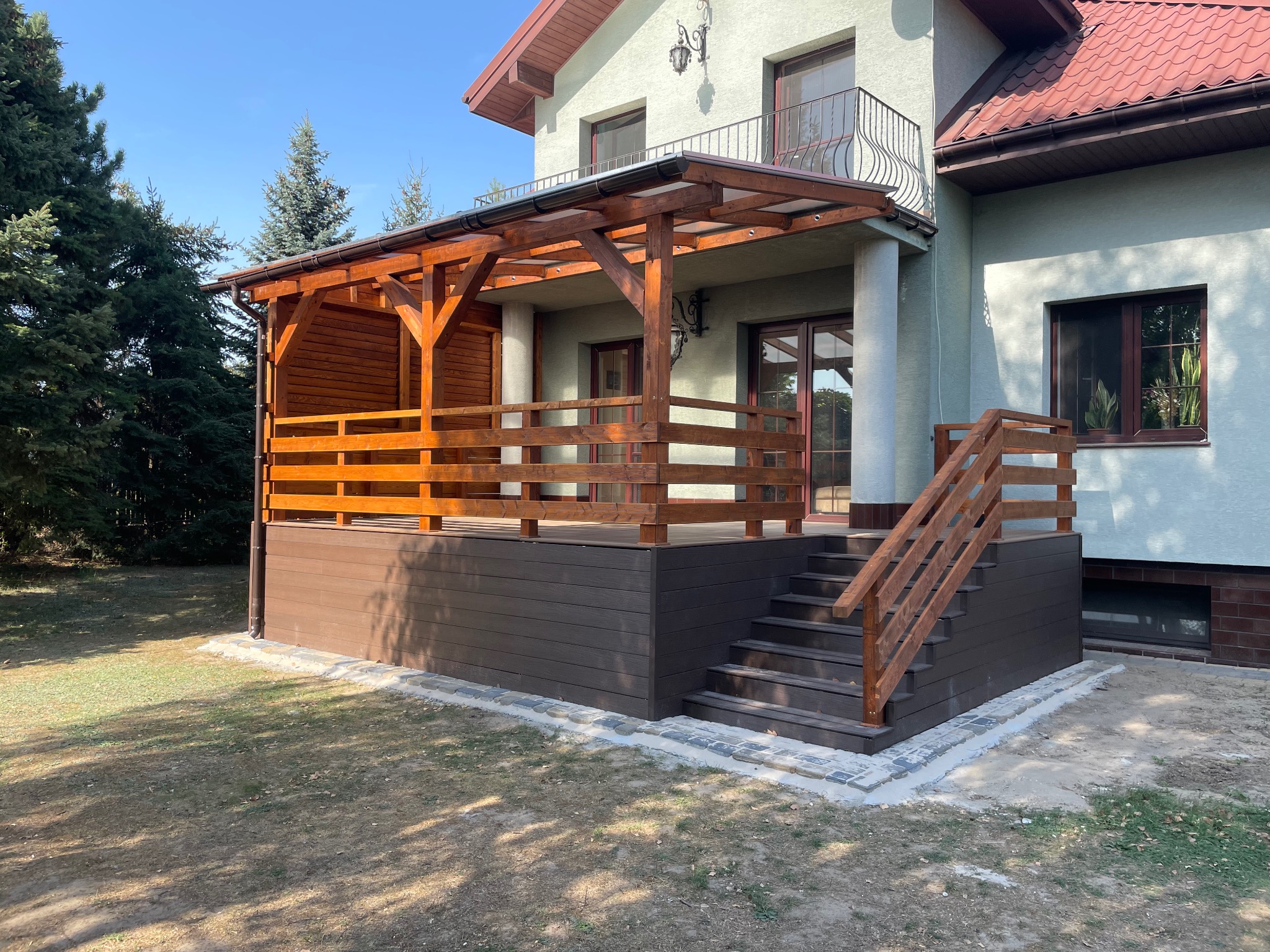 Zadaszony taras z brązowych desek kompozytowych z drewnianą balustradą i schodami, przylegający do jasnego domu z czerwonym dachem, otoczony zielenią.