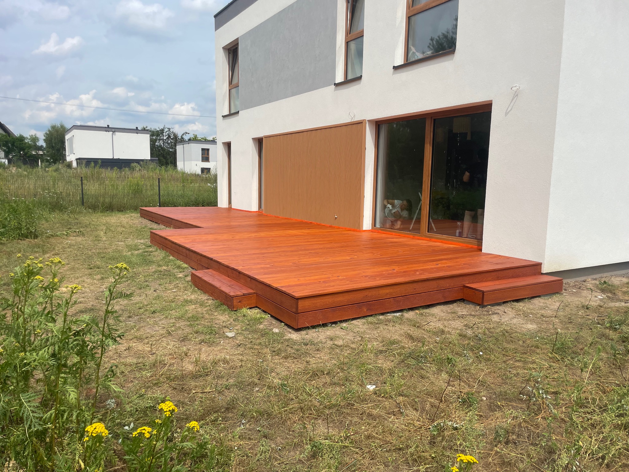 Nowoczesny dom z dużą, świeżo polakierowaną, drewnianą platformą tarasową z niskimi schodami, otoczony dziką roślinnością. Widoczne okna i elewacja domu w odcieniach bieli i szarości.