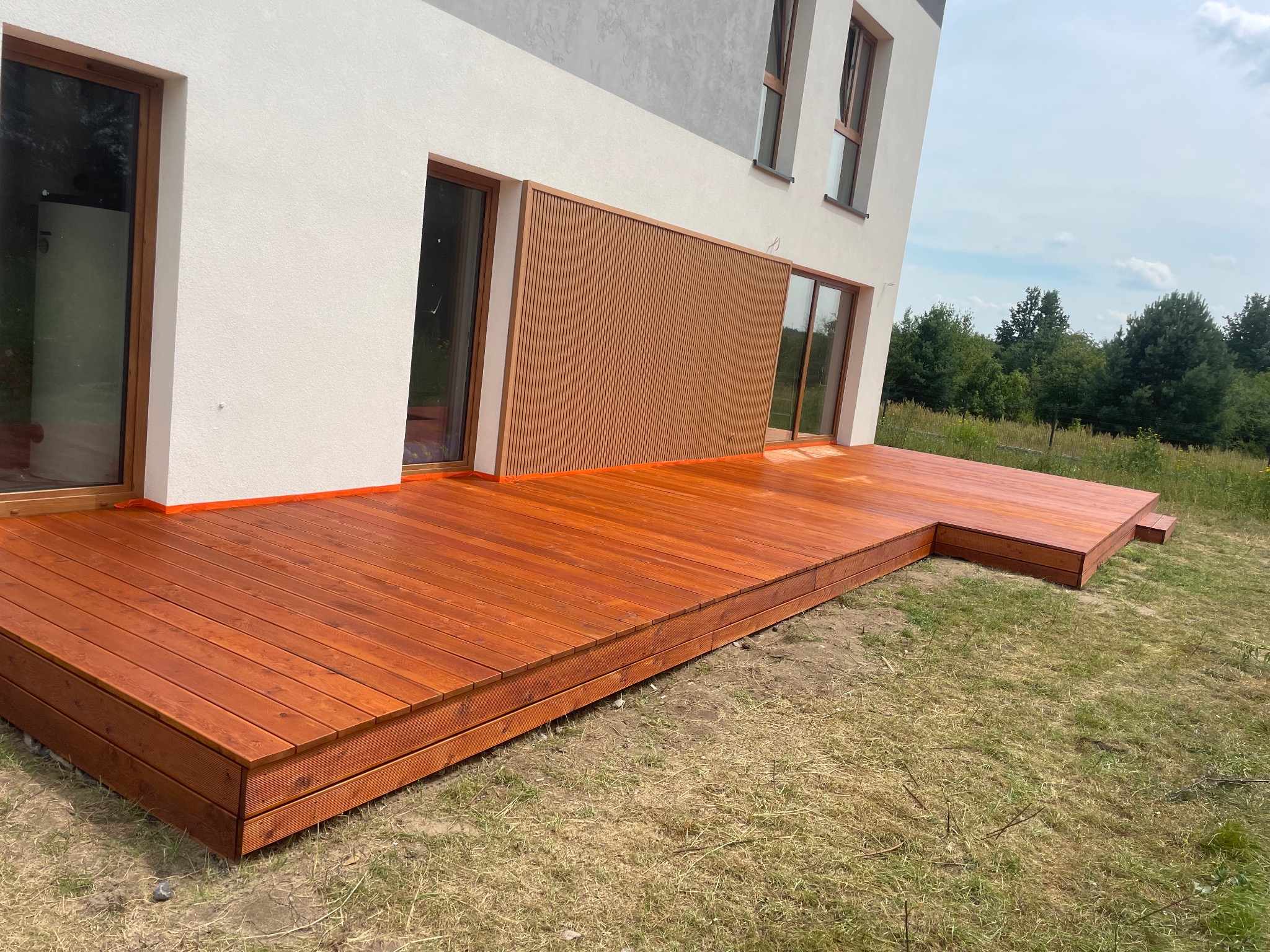 Nowoczesny dom z tarasem z drewna egzotycznego o ciepłym, brązowym odcieniu, z widoczną strukturą desek, obrzeżem zabezpieczonym pomarańczową taśmą malarską, otoczony trawnikiem i zielenią.