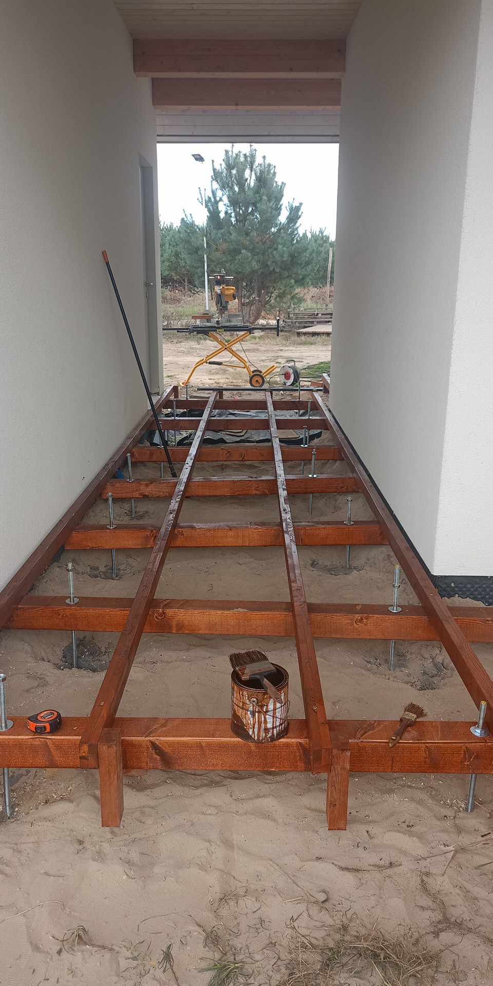 Konstrukcja nośna drewnianego tarasu w trakcie budowy, widoczne legary, słupki na metalowych kotwach, puszka z farbą i pędzle, w tle krajobraz z drzewami i sprzętem budowlanym.