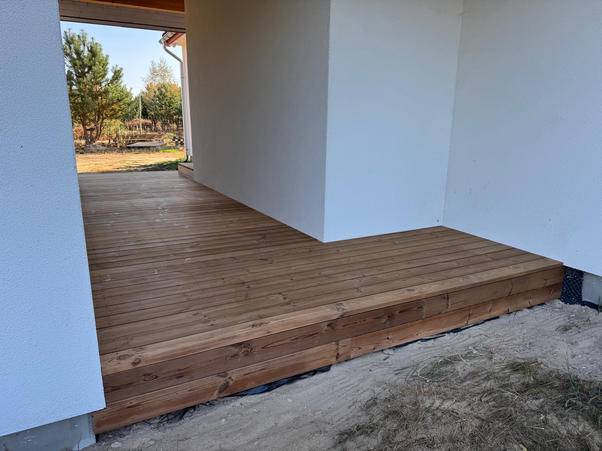 Nowo wybudowany taras z desek o ciepłym, brązowym odcieniu, przylegający do białej ściany budynku, widok na ogród z drzewami w tle.