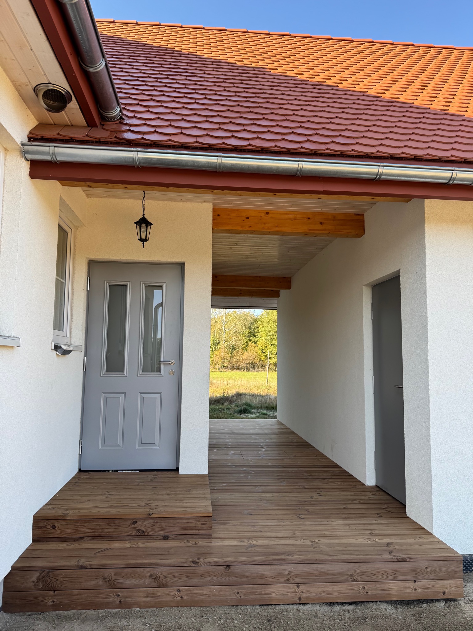 Zadaszone wejście do domu z drewnianym podestem i schodami, szare drzwi wejściowe z przeszkleniami, lampa zewnętrzna, widok na ogród w tle.