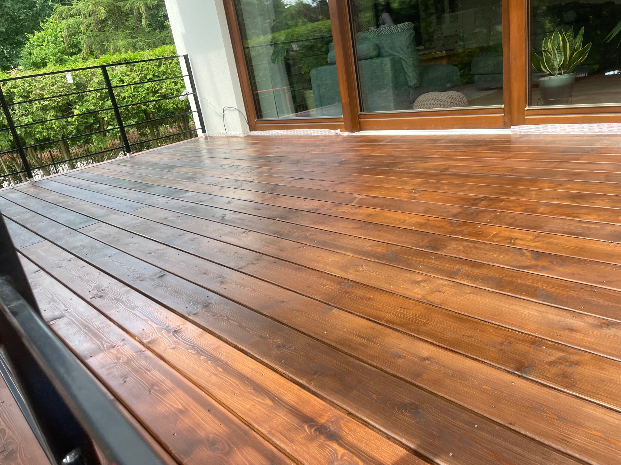 Świeżo polakierowany, drewniany taras z czarną, metalową balustradą i widokiem na zieleń, z dużymi oknami przesuwnymi w tle.