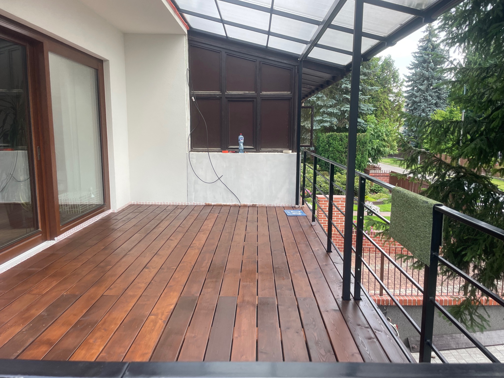 Zadaszony taras z drewnianą podłogą w trakcie budowy, widoczne narzędzia i materiały budowlane, czarna metalowa balustrada, widok na zieleń ogrodu