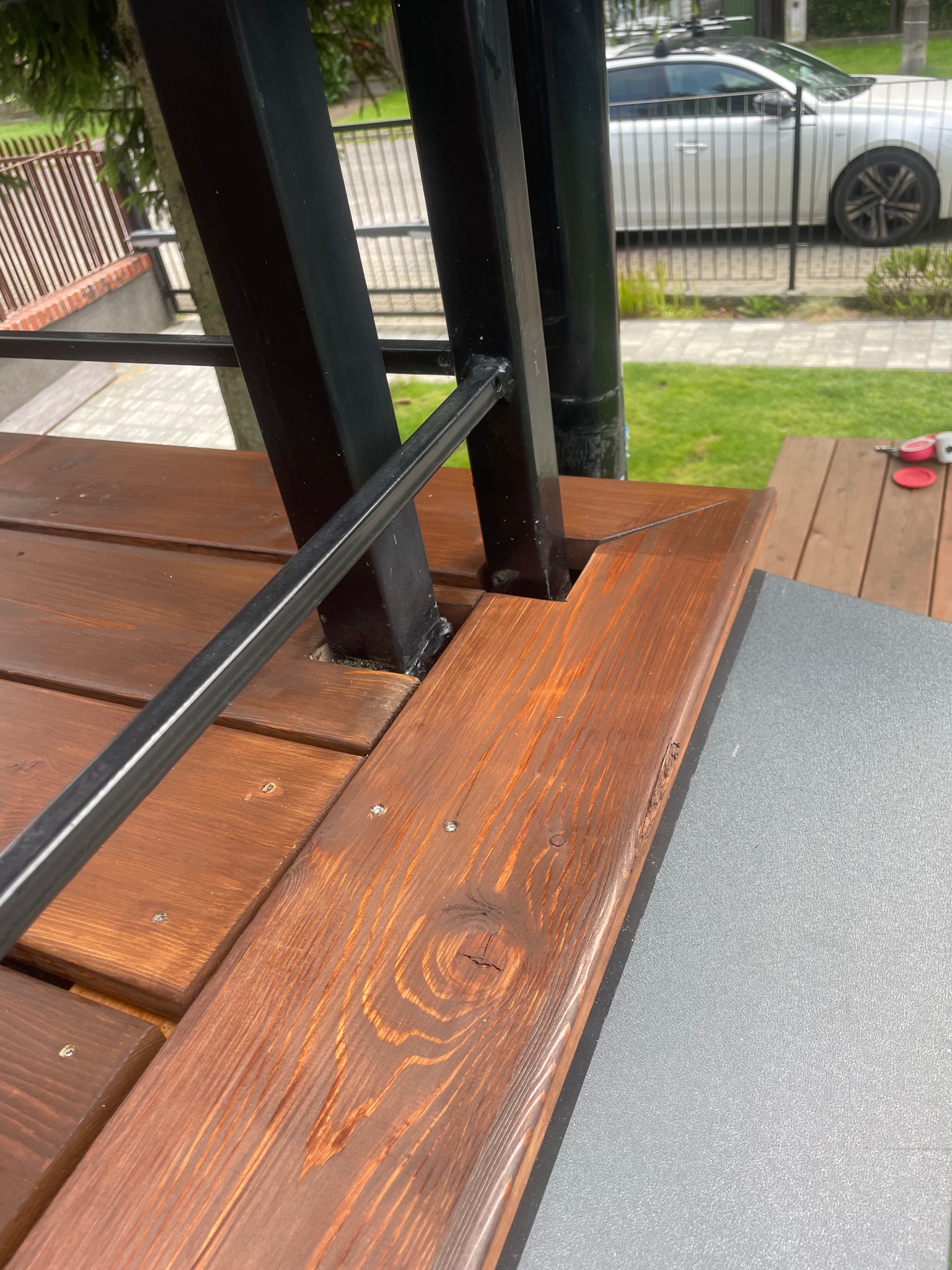 Fragment konstrukcji drewnianego tarasu z metalowymi elementami, widoczne łączenia desek i słupków, na drugim planie samochód