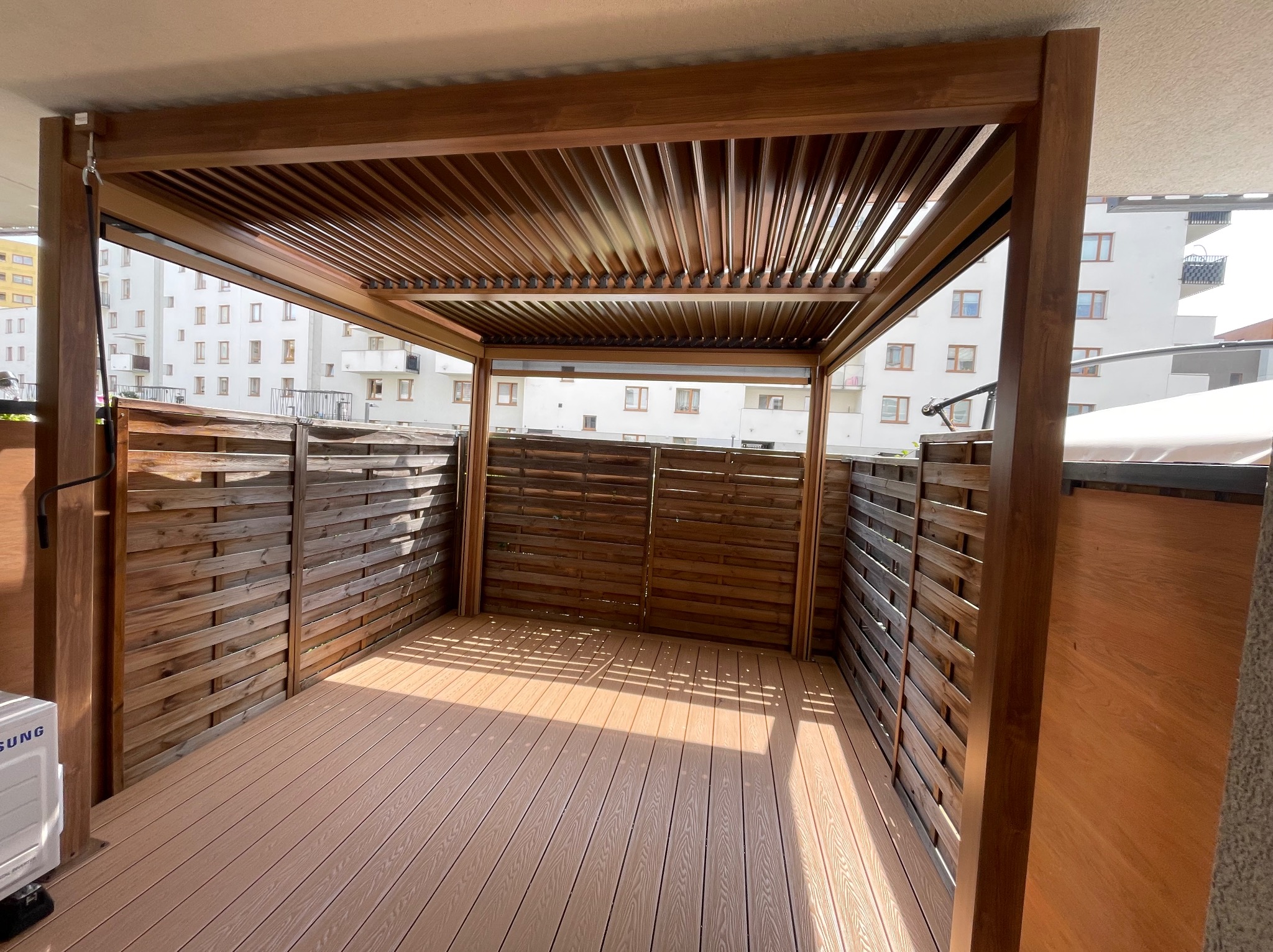 Zadaszona konstrukcja z drewna na tarasie w Warszawie. Drewniana podłoga i ażurowe ściany. Widok na budynki mieszkalne w tle. Nowoczesny design.