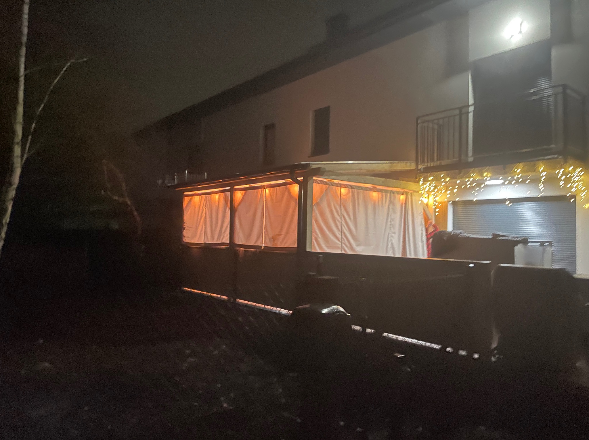 Oświetlona pergola drewniana z zasłonami na tle domu z balkonem i dekoracyjnymi lampkami, nocne ujęcie.