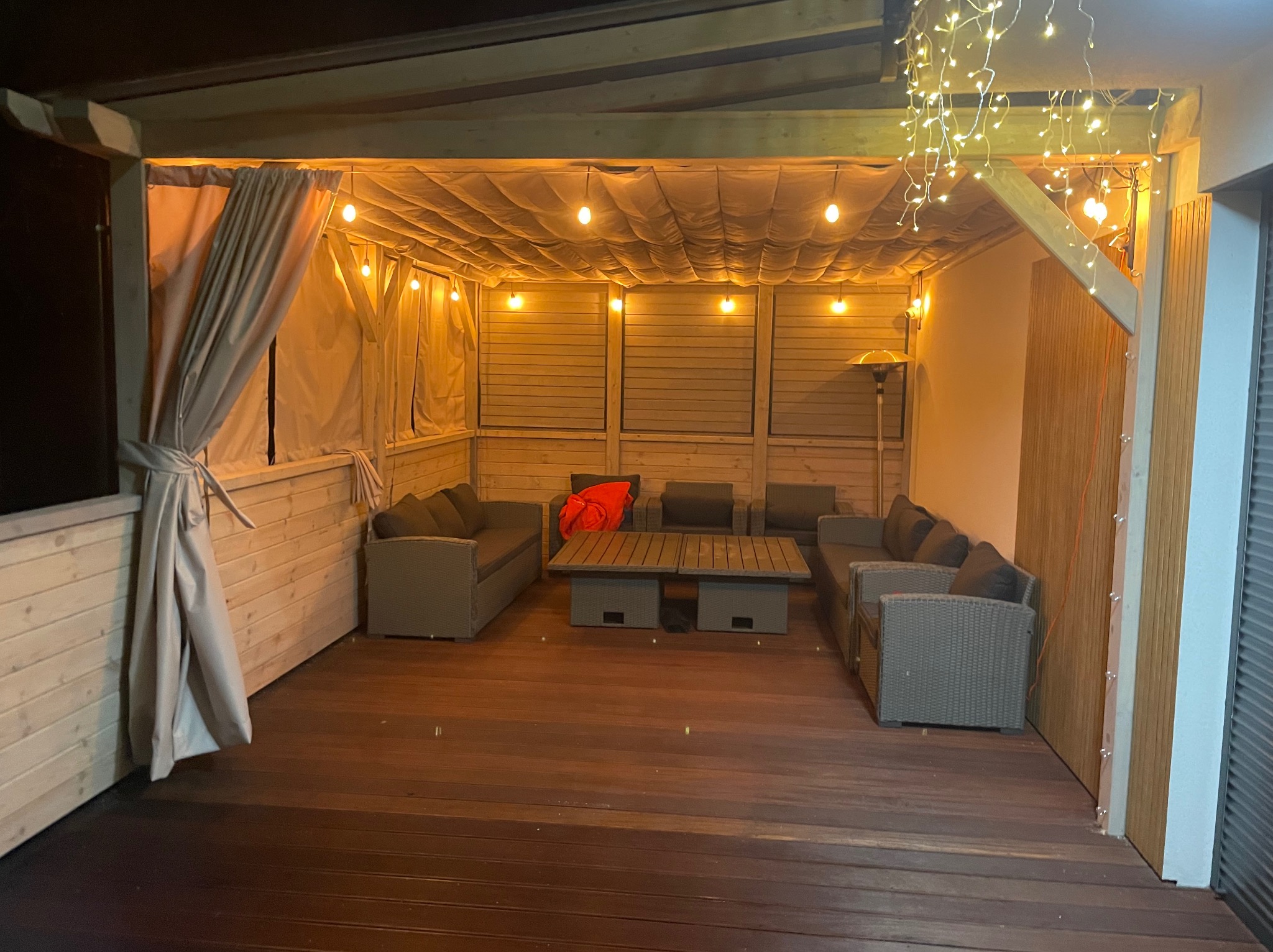 Zadaszony taras z drewnianą podłogą, oświetlony girlandami żarówek. Wnętrze wyposażone w meble ogrodowe i zasłony, tworzące przytulną strefę relaksu.