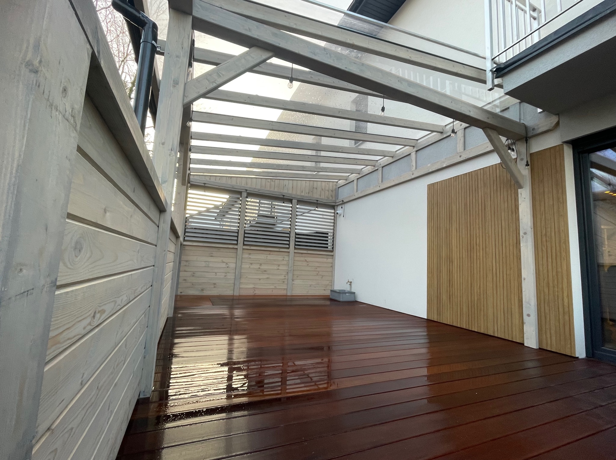 Zadaszony taras z drewnianą podłogą i szarą konstrukcją, oświetlony wiszącymi lampkami. Widok na fragment budynku i elementy wykończenia.