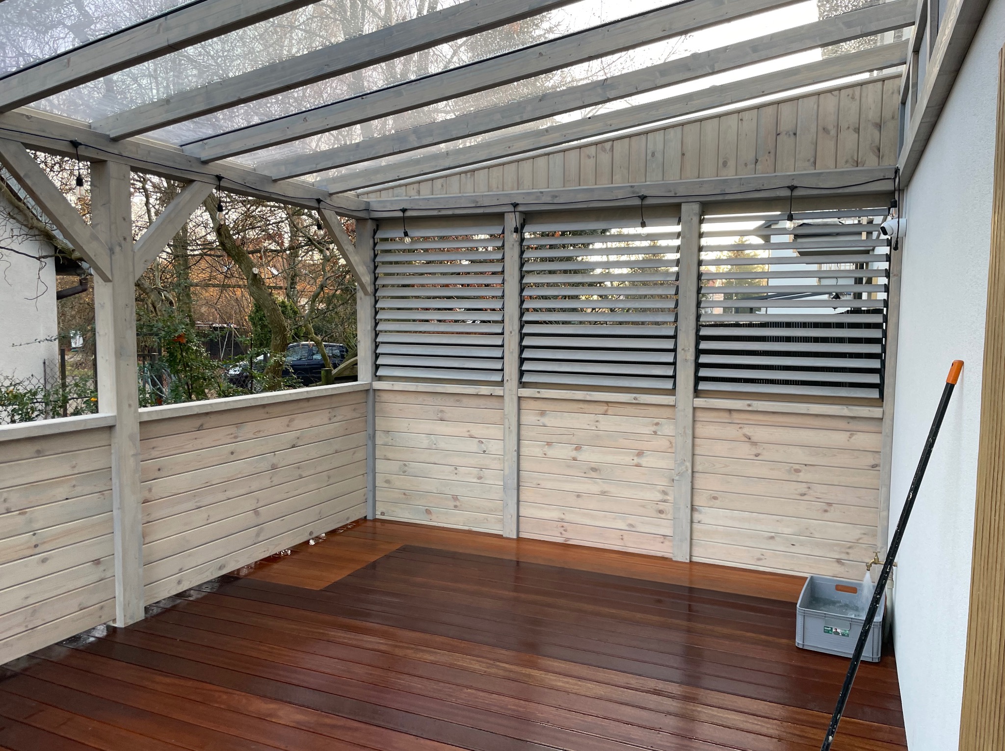 Zadaszony taras z drewnianą podłogą i ażurowymi panelami bocznymi, oświetlony girlandą żarówek. Widoczne drzewa w tle.