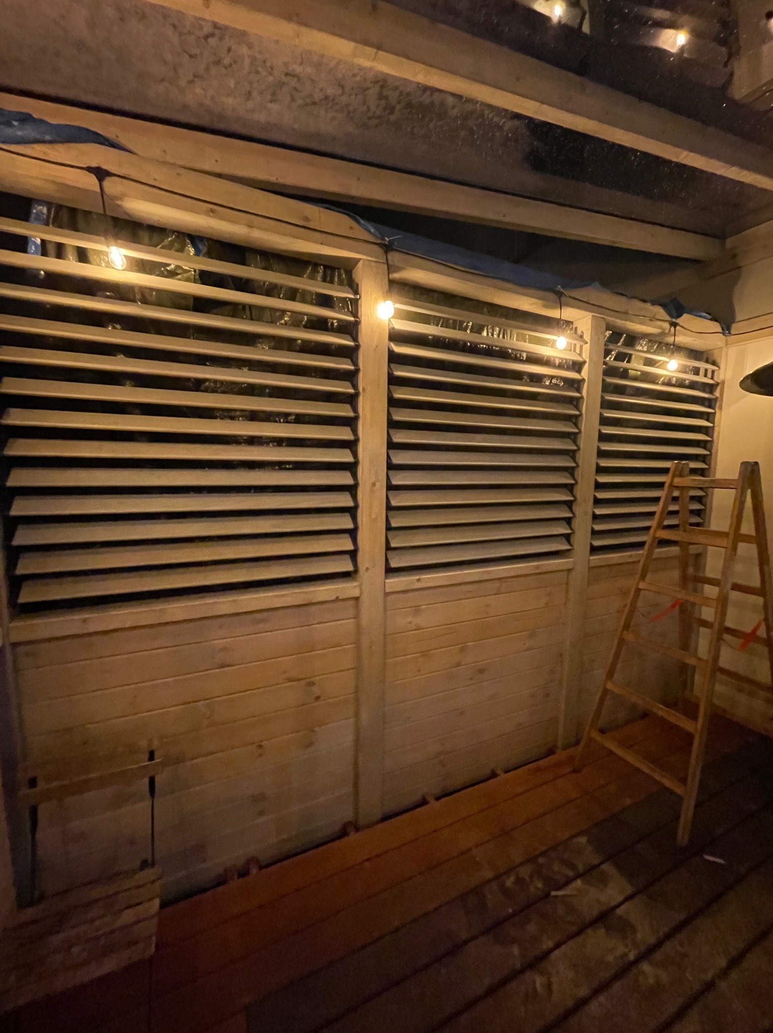 Drewniana konstrukcja z żaluzjami, oświetlona girlandą żarówek, z drewnianą podłogą i drabiną. Widoczne elementy montażowe i folia zabezpieczająca.