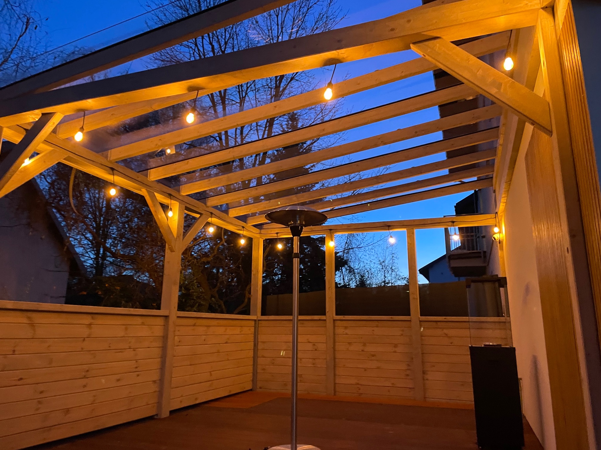 Zadaszona konstrukcja z drewna, oświetlona girlandą żarówek, z widocznym grzejnikiem stojącym i elementami zabudowy, na tle wieczornego nieba.