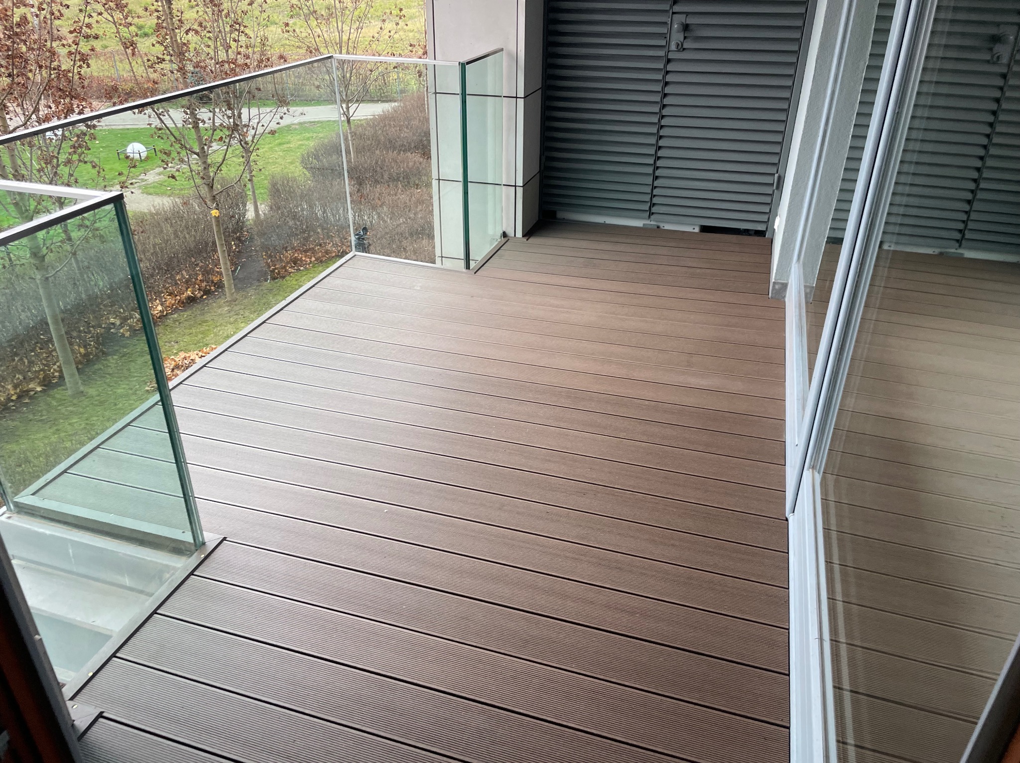 Nowoczesny balkon z brązową podłogą z desek kompozytowych i szklanymi balustradami, widok na zieleń. Minimalistyczny design, jasne otoczenie.