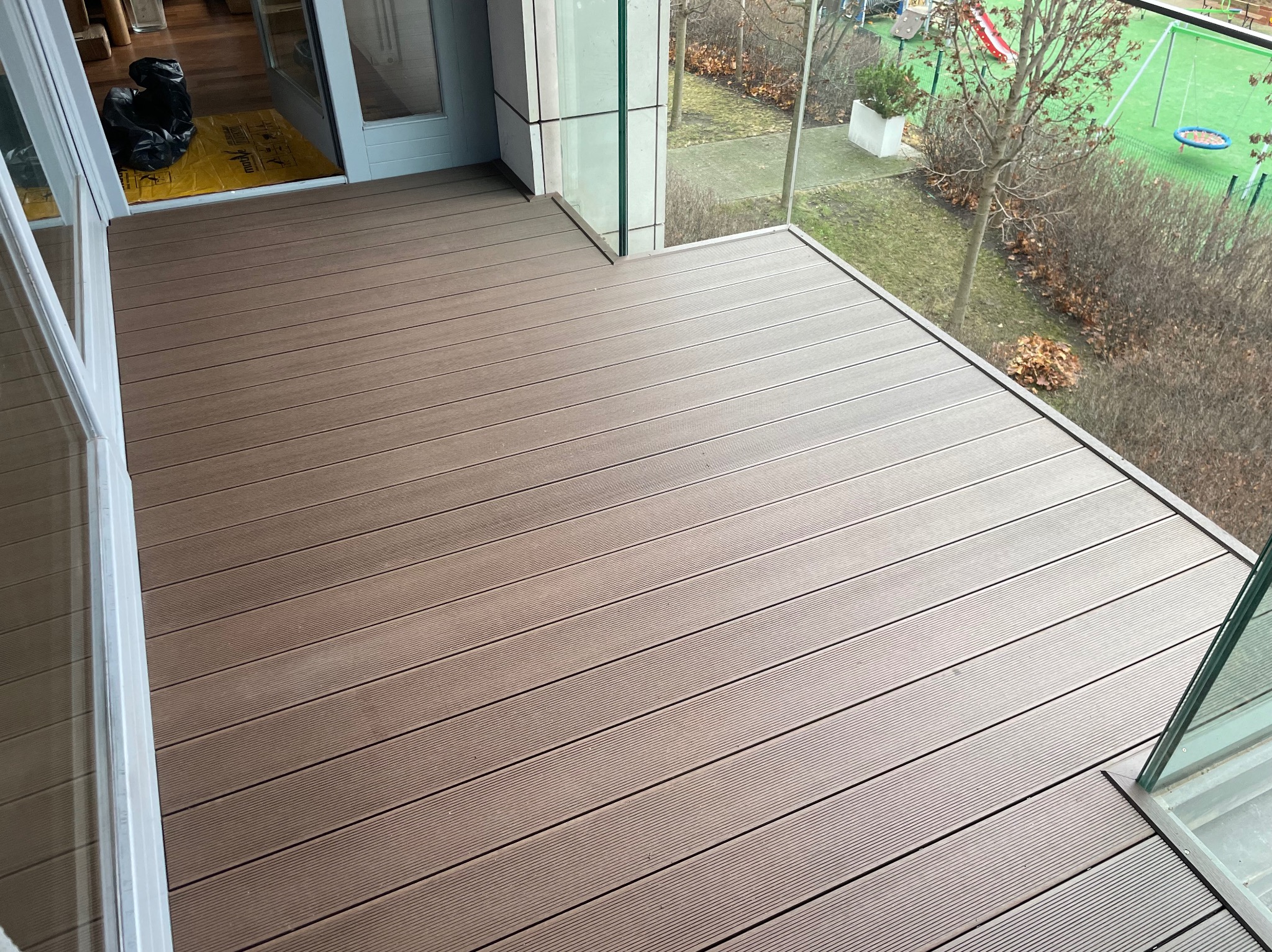 Nowoczesny taras kompozytowy z widokiem na plac zabaw, zabezpieczony szklaną balustradą. Brązowe deski ułożone równolegle, widoczne detale wykończenia.