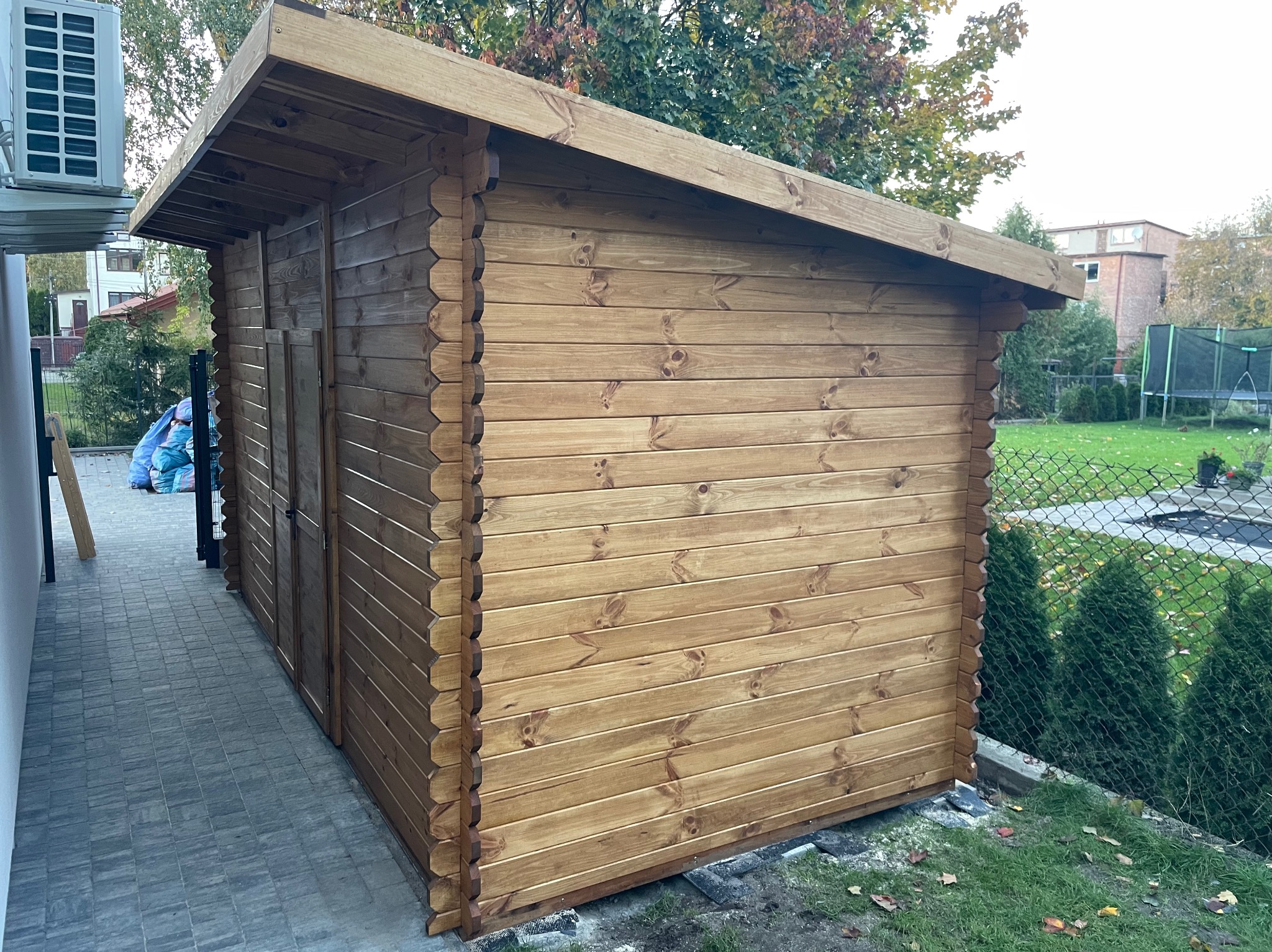 Drewniana konstrukcja z bali, prawdopodobnie domek narzędziowy, z widocznym detalem łączenia bali i zadaszeniem, na tle zieleni i kostki brukowej.