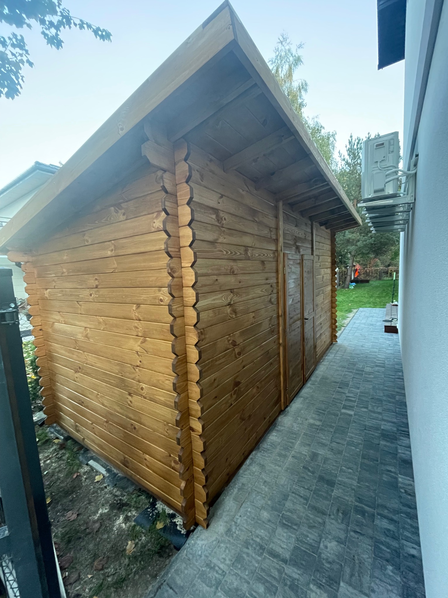 Drewniana altana z dwuspadowym dachem, wykonana z bali, zamykana na drzwi, ustawiona na kostce brukowej, przylegająca do białej ściany budynku.