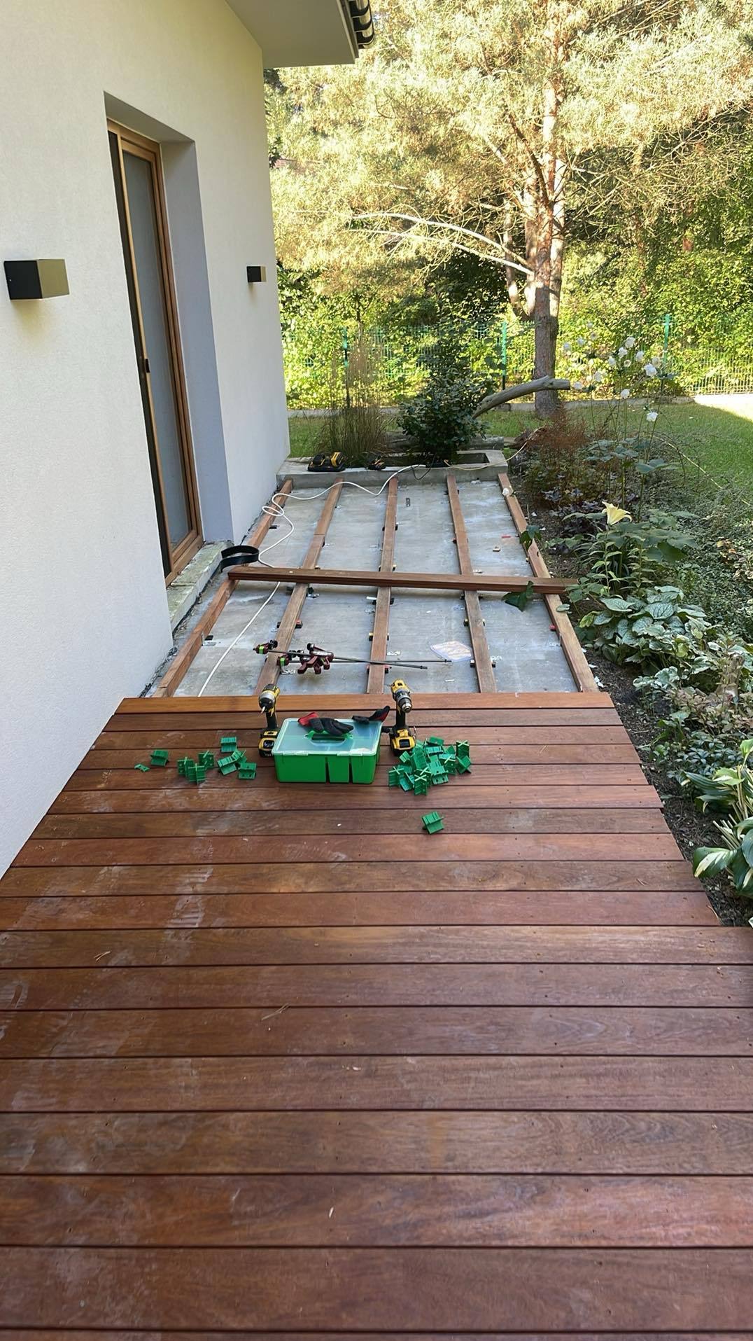 Montaż brązowego tarasu z desek przy domu. Widoczne narzędzia, poziomica i elementy konstrukcyjne. Część tarasu już ułożona, reszta w trakcie budowy.