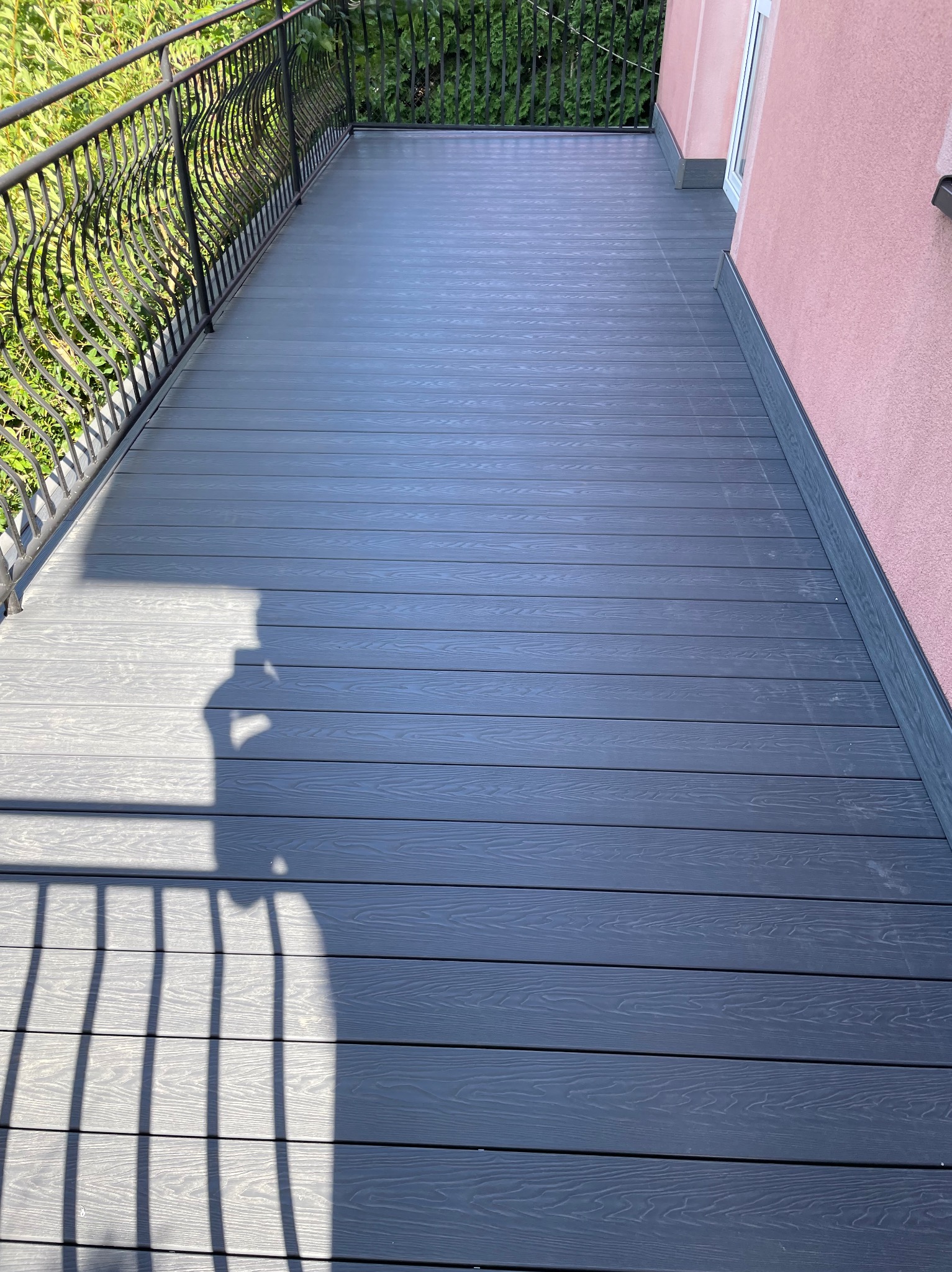 Szary taras kompozytowy z ozdobną, metalową balustradą i cieniem osoby robiącej zdjęcie, przylegający do różowej ściany budynku.