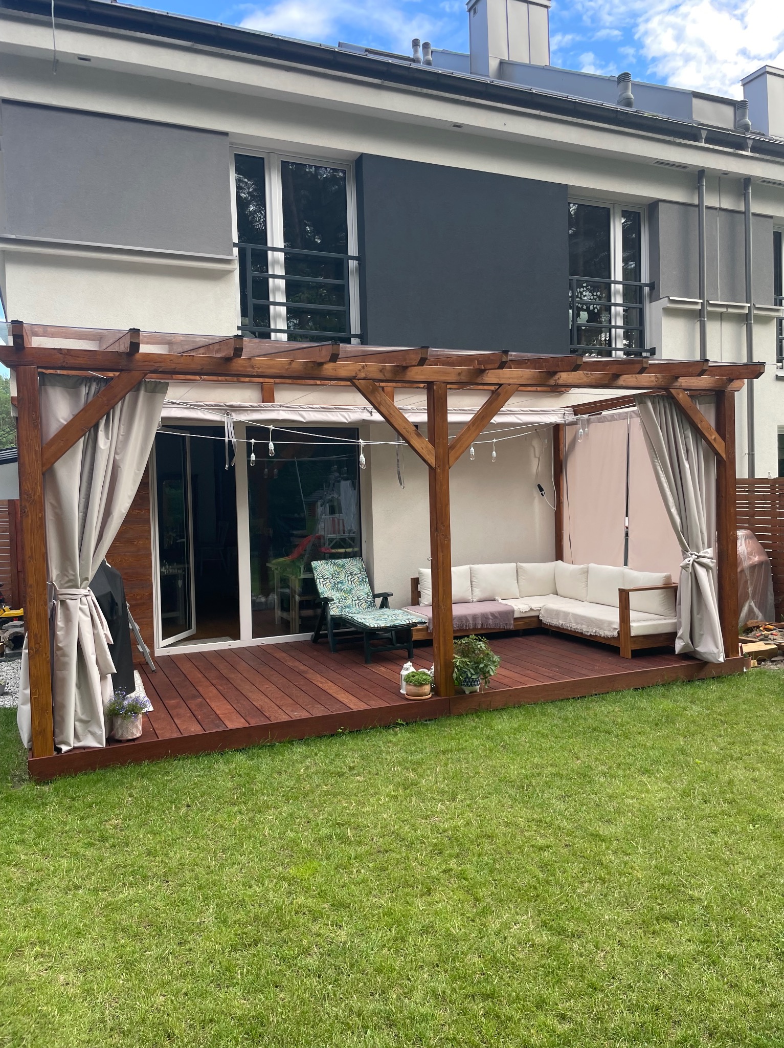 Zadaszony taras z drewnianą pergolą i zasłonami, wyposażony w meble ogrodowe, z widokiem na trawnik i nowoczesny dom.