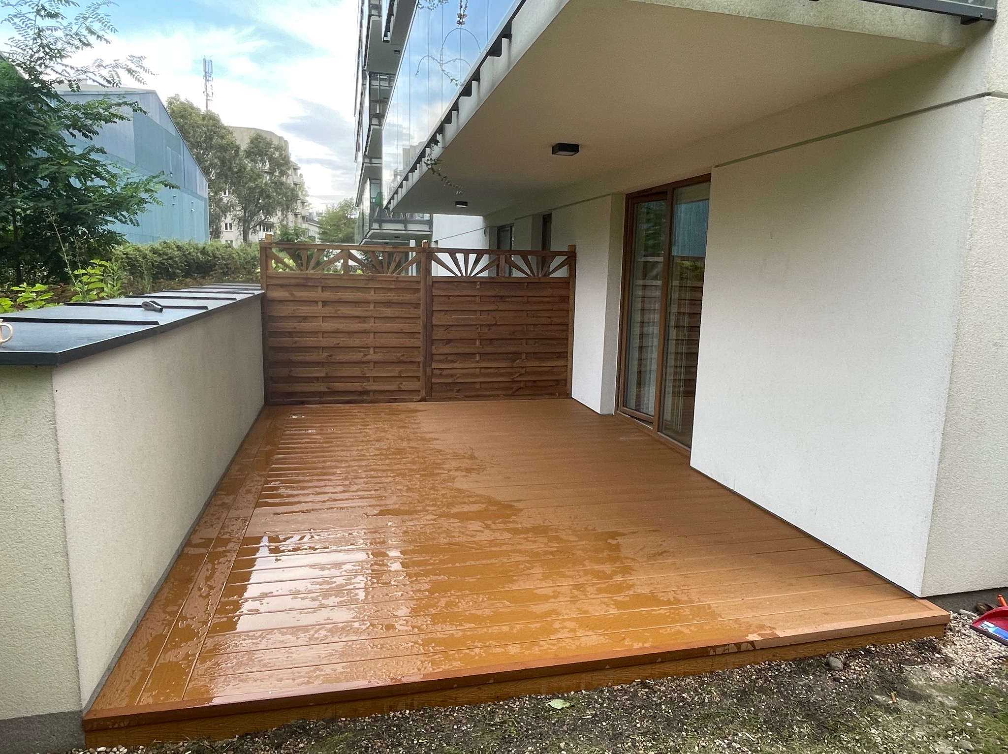 Nowoczesny taras na balkonie z drewnianą podłogą i ażurową balustradą, widok z góry, po deszczu. Minimalistyczny design, jasne ściany budynku w tle.