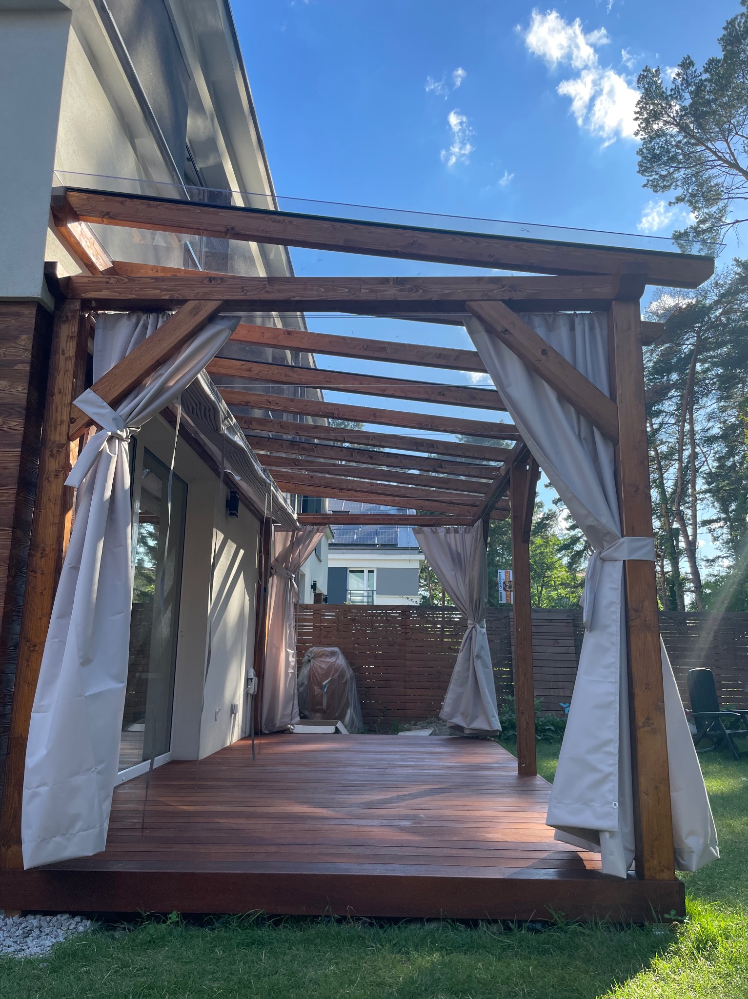 Drewniana pergola z przezroczystym zadaszeniem i jasnymi zasłonami, przylegająca do domu. Widoczny fragment tarasu z desek i zadbany trawnik. Słoneczny dzień.
