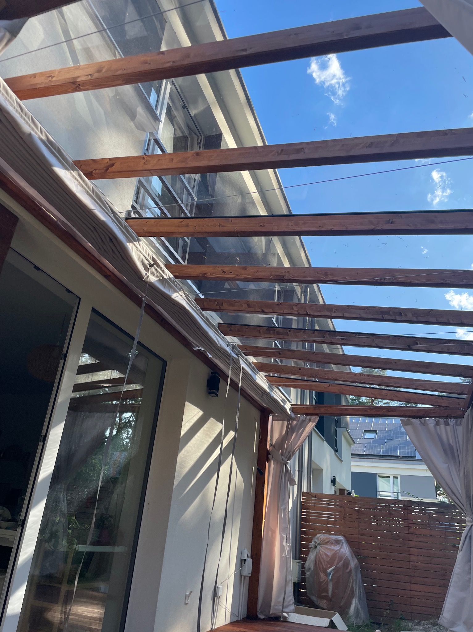 Zadaszenie tarasu z drewnianą konstrukcją i przezroczystym dachem, widok na błękitne niebo. Zasłony po bokach dodają prywatności. Nowoczesny design.