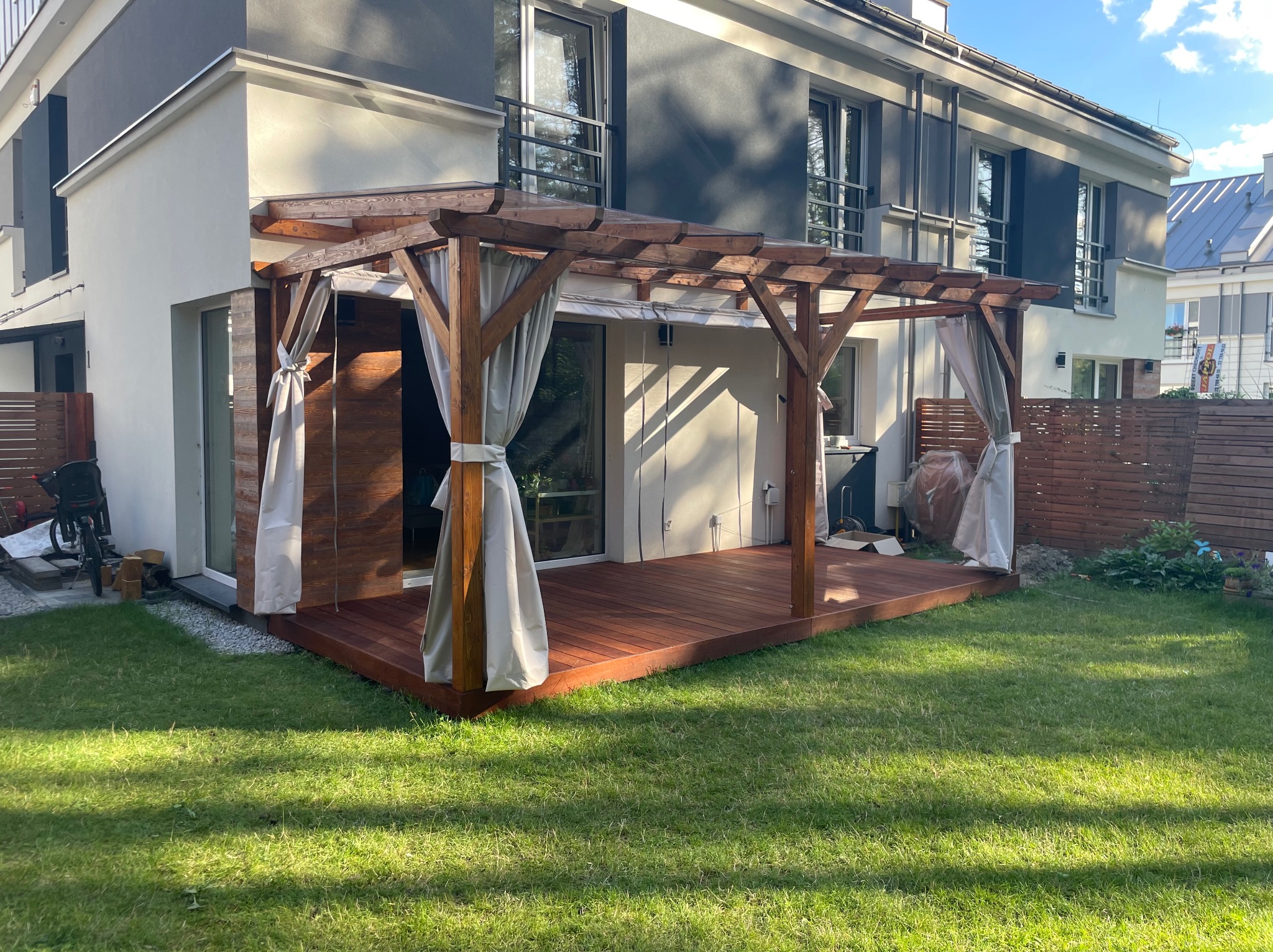 Zadaszony, drewniany taras z zasłonami przy nowoczesnym domu. Ciemne deski, jasne zasłony, drewniana konstrukcja zadaszenia. Zielony trawnik.