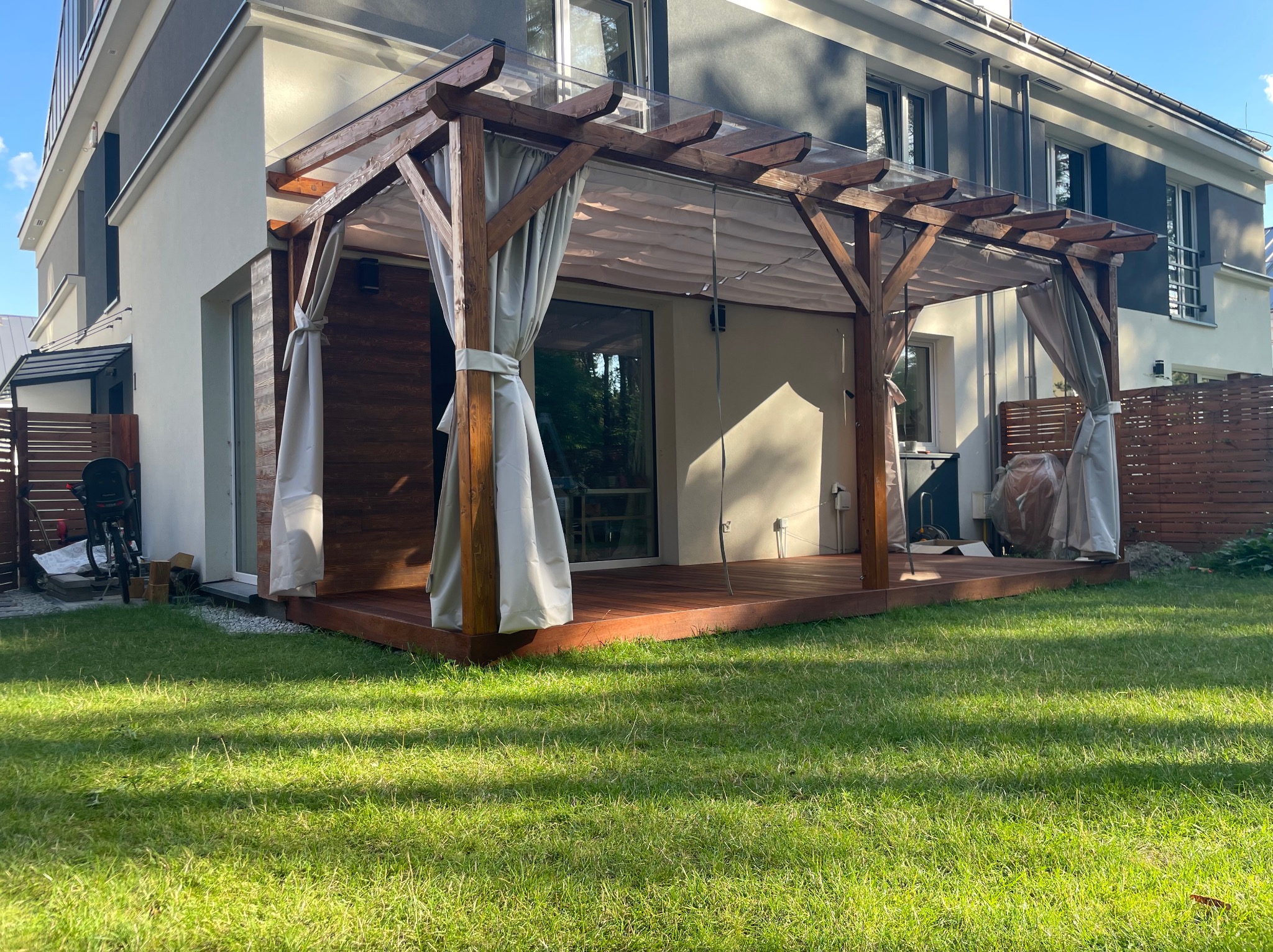 Drewniana pergola z jasnymi zasłonami, przylegająca do nowoczesnego domu. Widoczny drewniany taras i zadbany trawnik. Całość w słoneczny dzień.