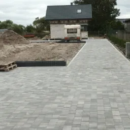 Ułożona szara kostka brukowa na podjeździe, w tle budynek w trakcie budowy i przyczepa kempingowa.