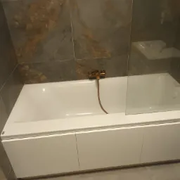 Biała wanna z zabudową szafkową i złotą armaturą na tle ciemnych, marmurkowych płytek w łazience.