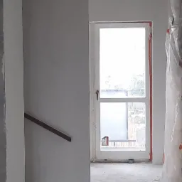 Wnętrze w trakcie remontu: widoczne surowe ściany przygotowane do malowania, zabezpieczone okno folią malarską, oraz fragment schodów z drewnianą poręczą. W tle widoczne elementy instalacji...