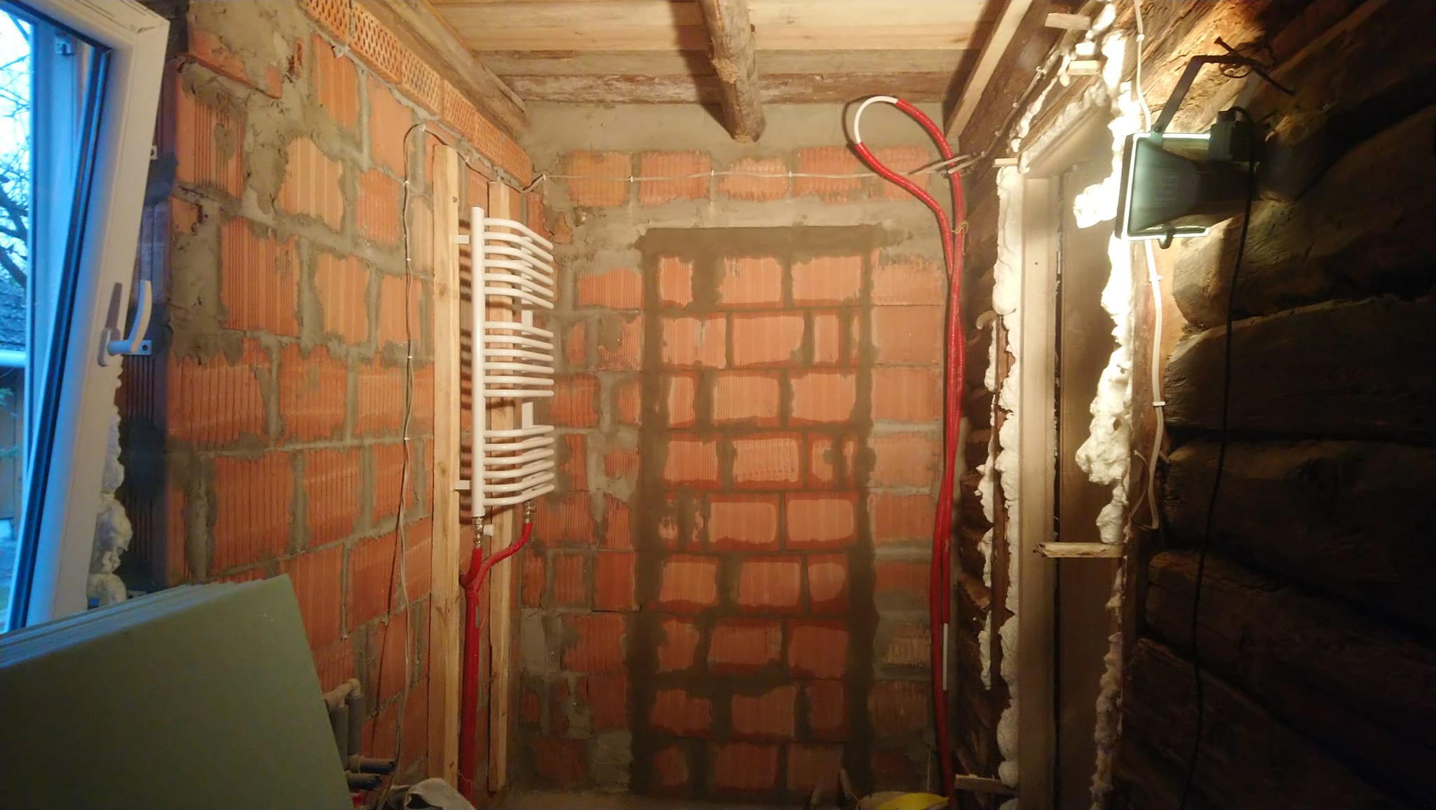 Wnętrze pomieszczenia w trakcie prac murarskich z widocznymi cegłami, instalacją grzewczą i oknem z widokiem na zewnątrz, oraz fragmentem ściany z drewnianych bali zabezpieczonej pianką montażową.