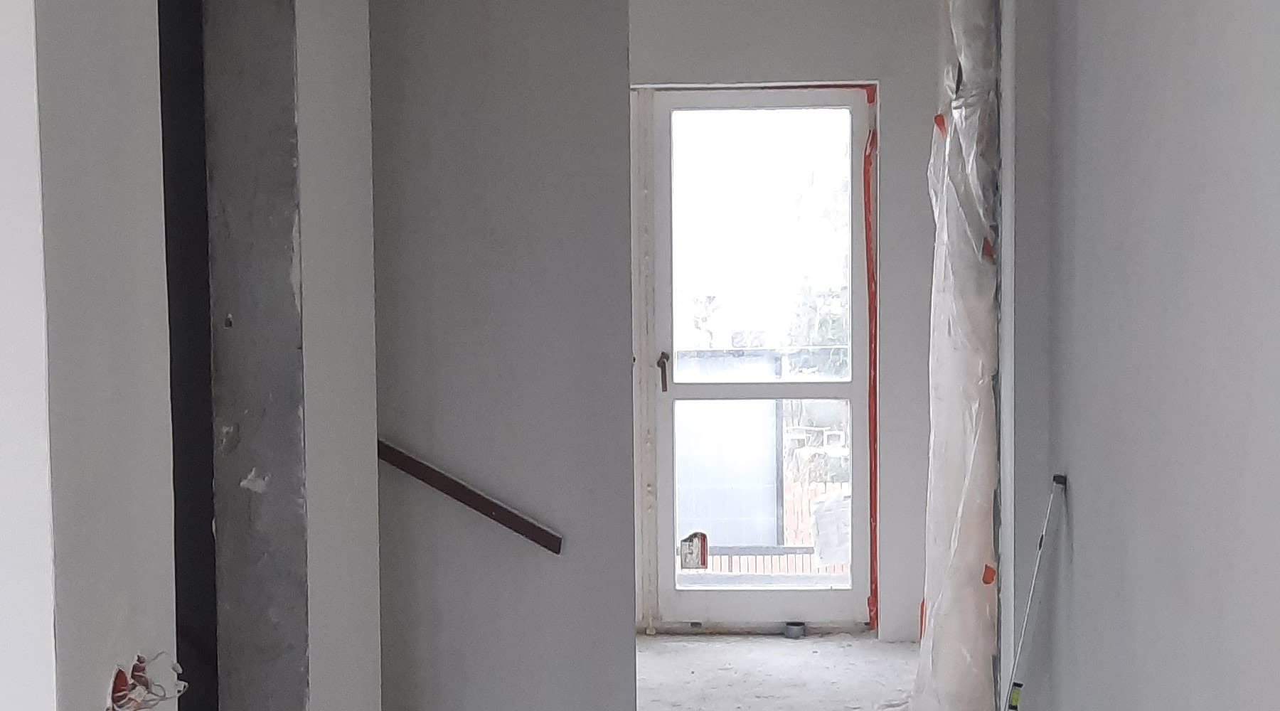 Wnętrze w trakcie remontu: widoczne surowe ściany przygotowane do malowania, zabezpieczone okno folią malarską, oraz fragment schodów z drewnianą poręczą. W tle widoczne elementy instalacji...