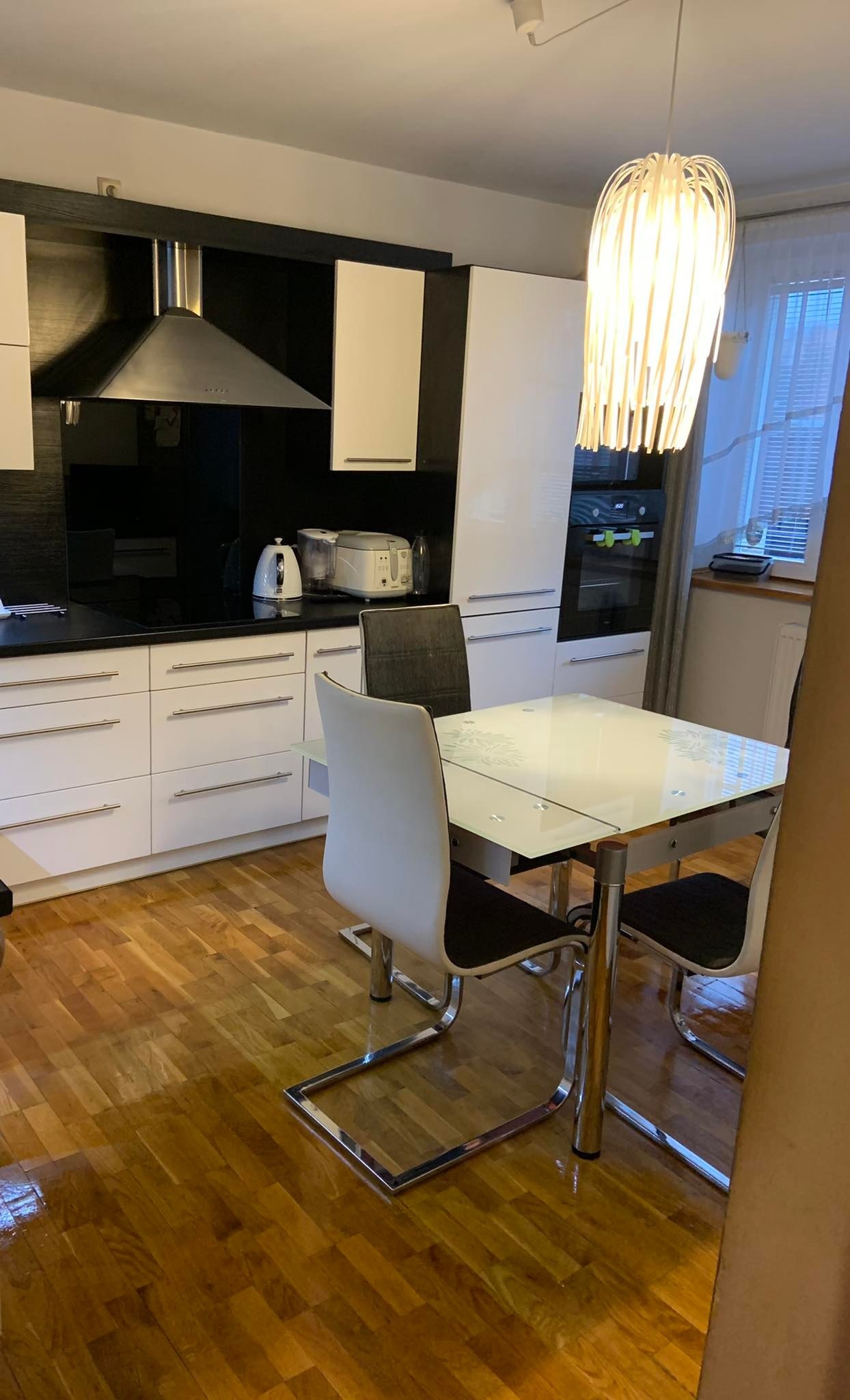 Jasna kuchnia z białymi szafkami, czarnym panelem nad blatem, szklanym stołem z motywem liści i nowoczesną lampą wiszącą.