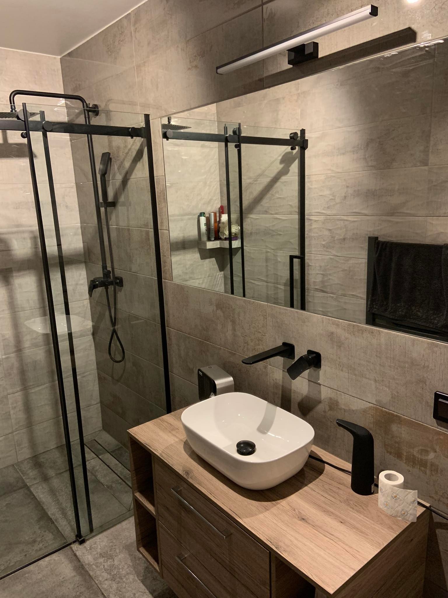 Nowoczesna łazienka z kabiną prysznicową typu walk-in, umywalką nablatową i czarną armaturą na tle płytek imitujących beton.