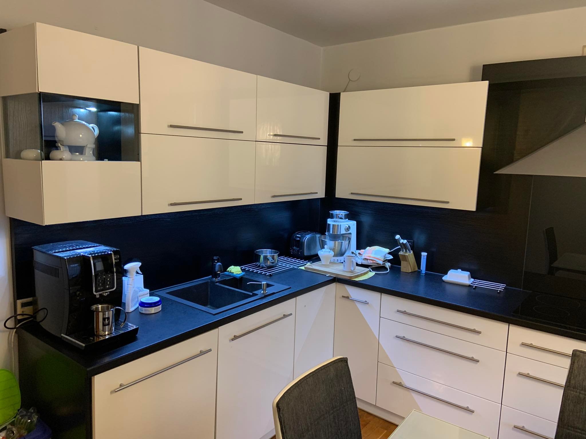 Nowoczesna, biała kuchnia z czarnym blatem, wyposażona w ekspres do kawy, czajnik i inne akcesoria kuchenne, widok na umywalkę i fragment jadalni z krzesłem.